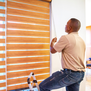 Man installing beige window blinds on a bright window using a stepladder