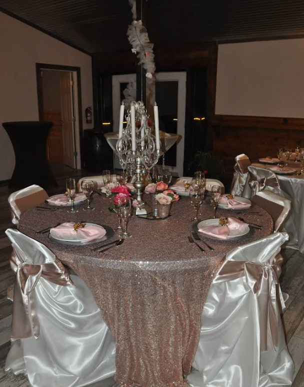 Round table set for a formal event, covered in rose gold sequins. Silverware, pink napkins, floral centerpiece.