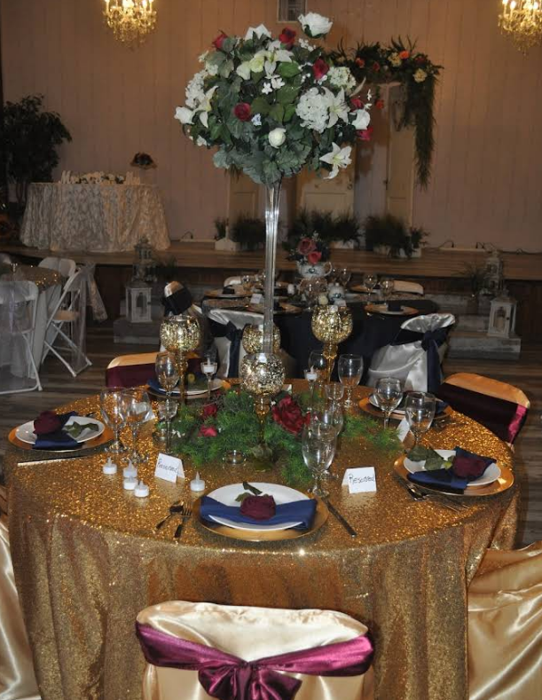 Wedding reception table setting with gold sequin tablecloth, floral centerpiece, and burgundy accents.