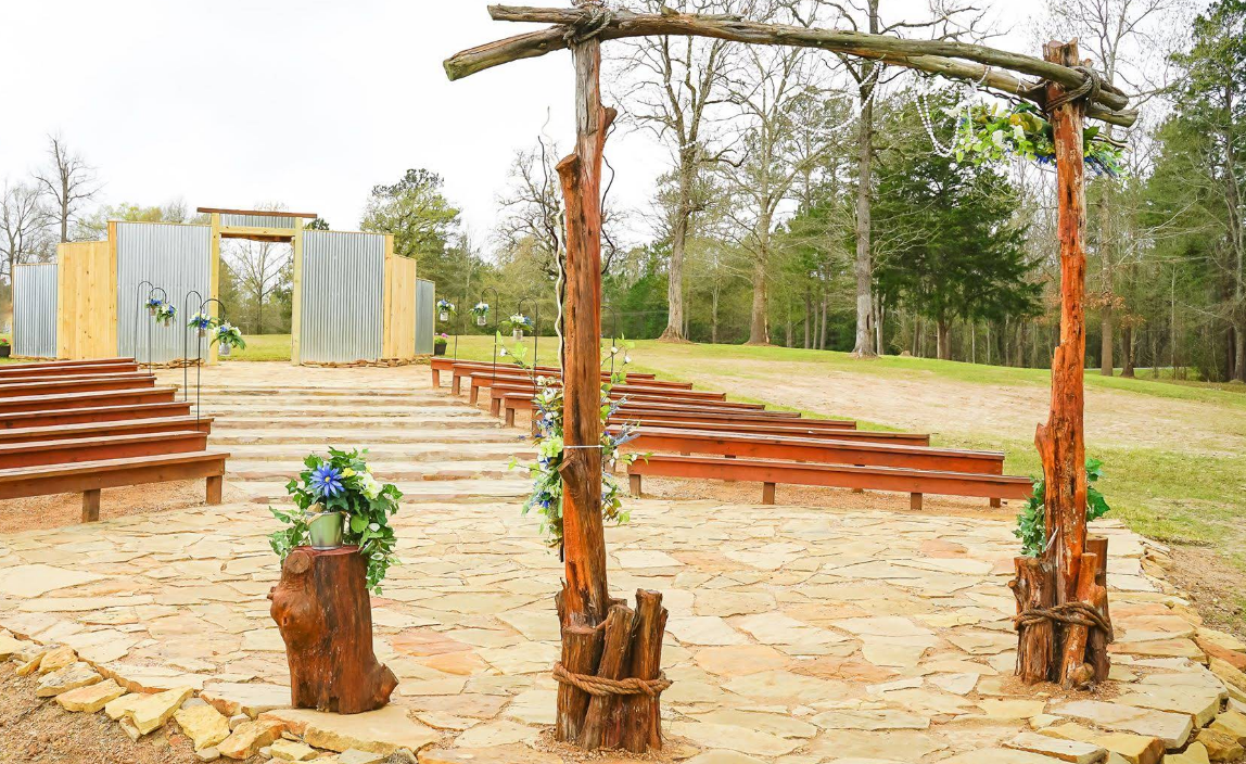 Outdoor wedding venue with rustic arch, benches, and stage, surrounded by trees.