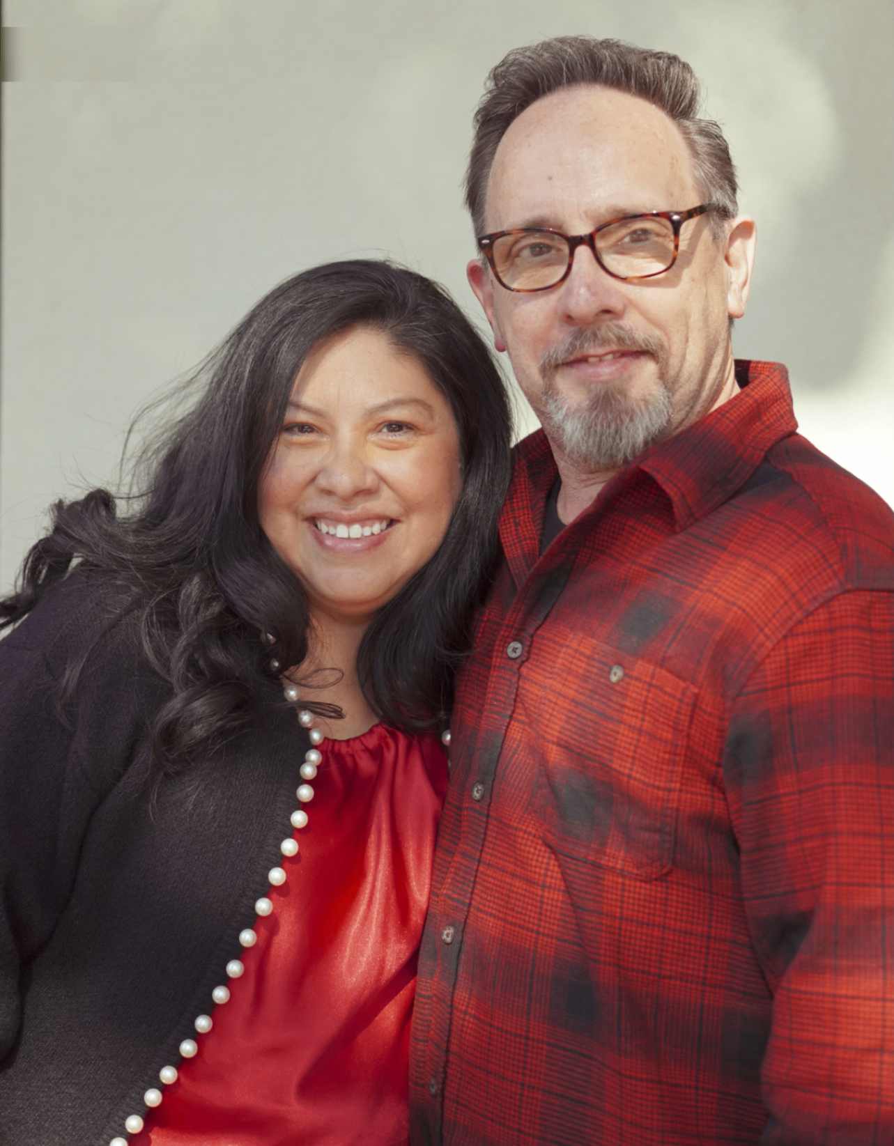 Woman and man smiling, posing together. Woman in red top, black sweater. Man in red plaid shirt, glasses.
