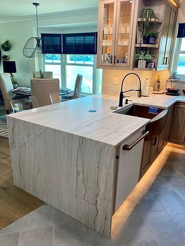 Kitchen island with light-colored, patterned countertop and cabinetry. Black faucet, under-cabinet lighting.