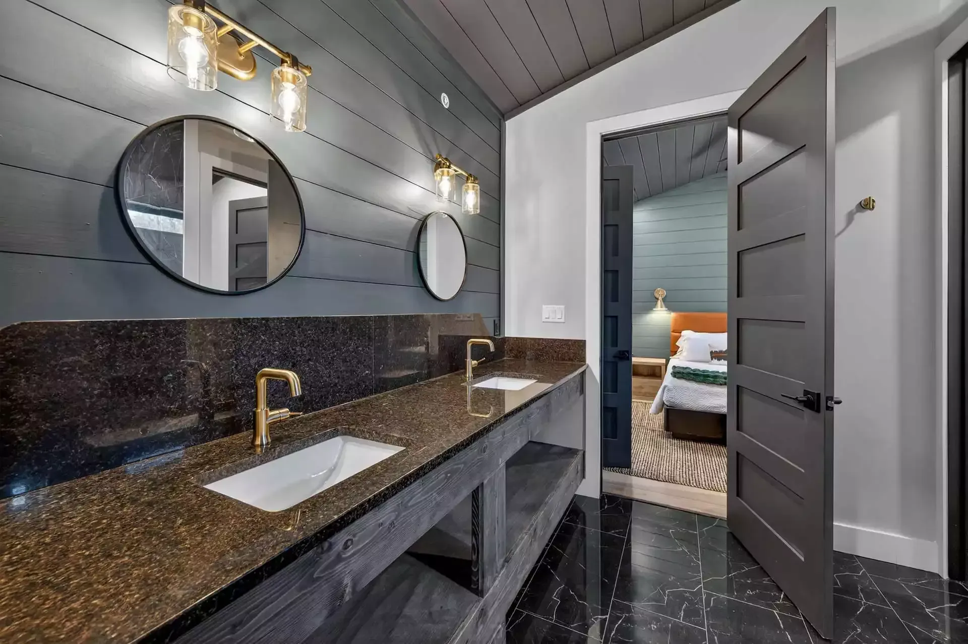 Modern bathroom with dark marble vanity, round mirrors, and open door to a bedroom with a bed