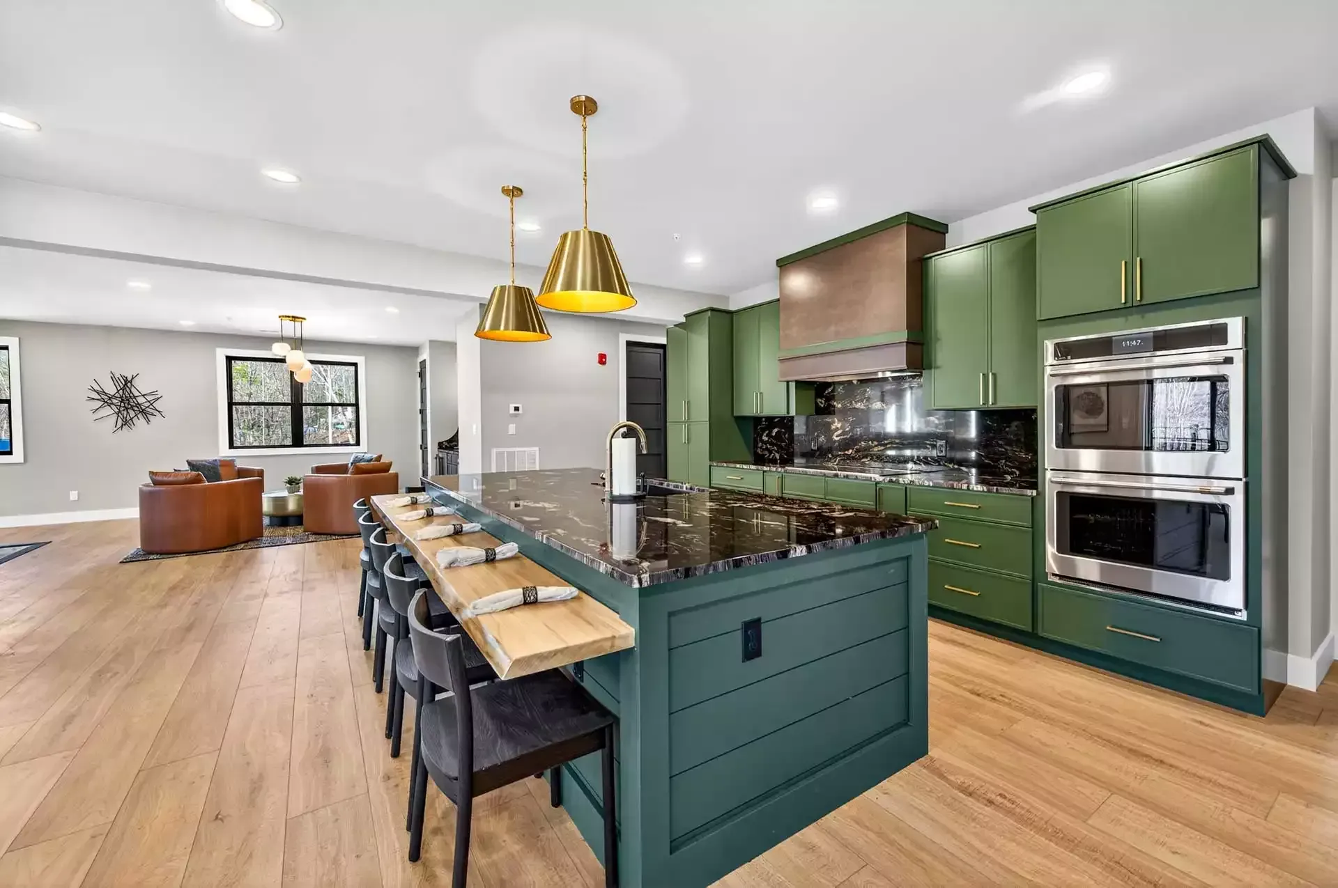 Bright open kitchen with green cabinets, black island, gold pendant lights, and wood floors opening to a dining area