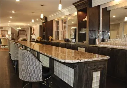 Long bar with granite countertop and upholstered chairs in a dimly lit room with mirrors and liquor display.