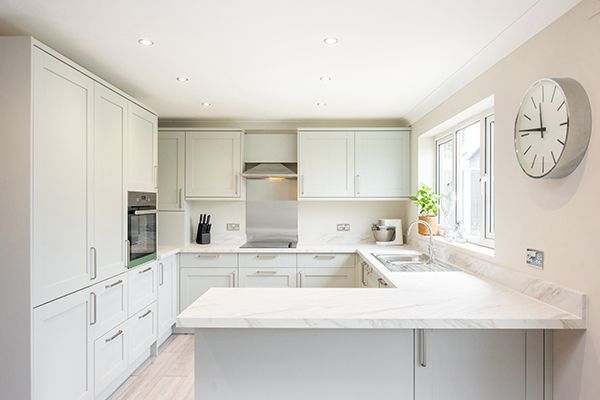 Modern white kitchen with marble kitchen countertops and built-in cabinets near window. U-shaped kitchen with light gray cabinets, white countertops, and a window with a clock.