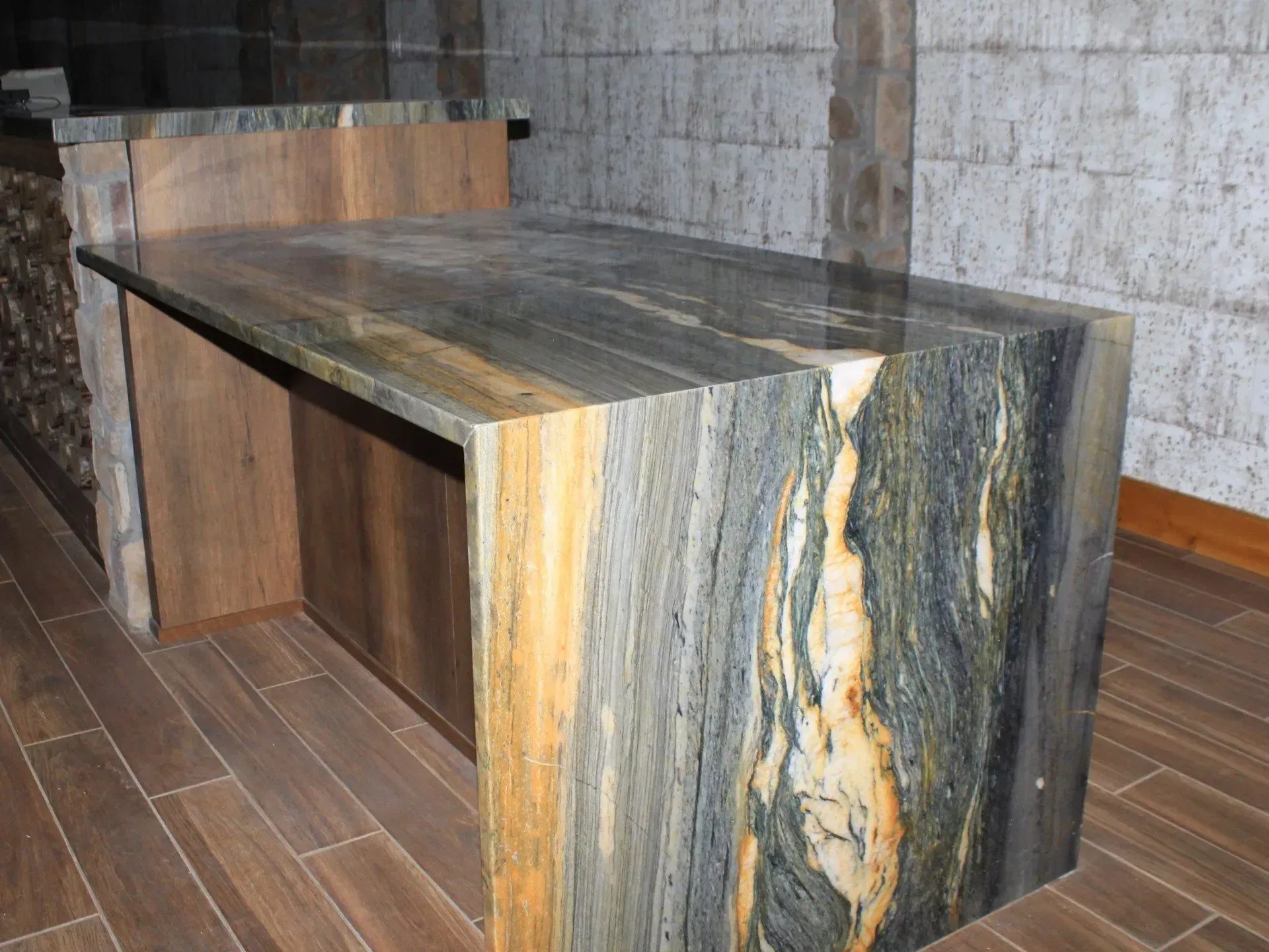 Countertop with gray, gold, and blue stone, with light wood cabinet. Brown floor, rough stone wall.