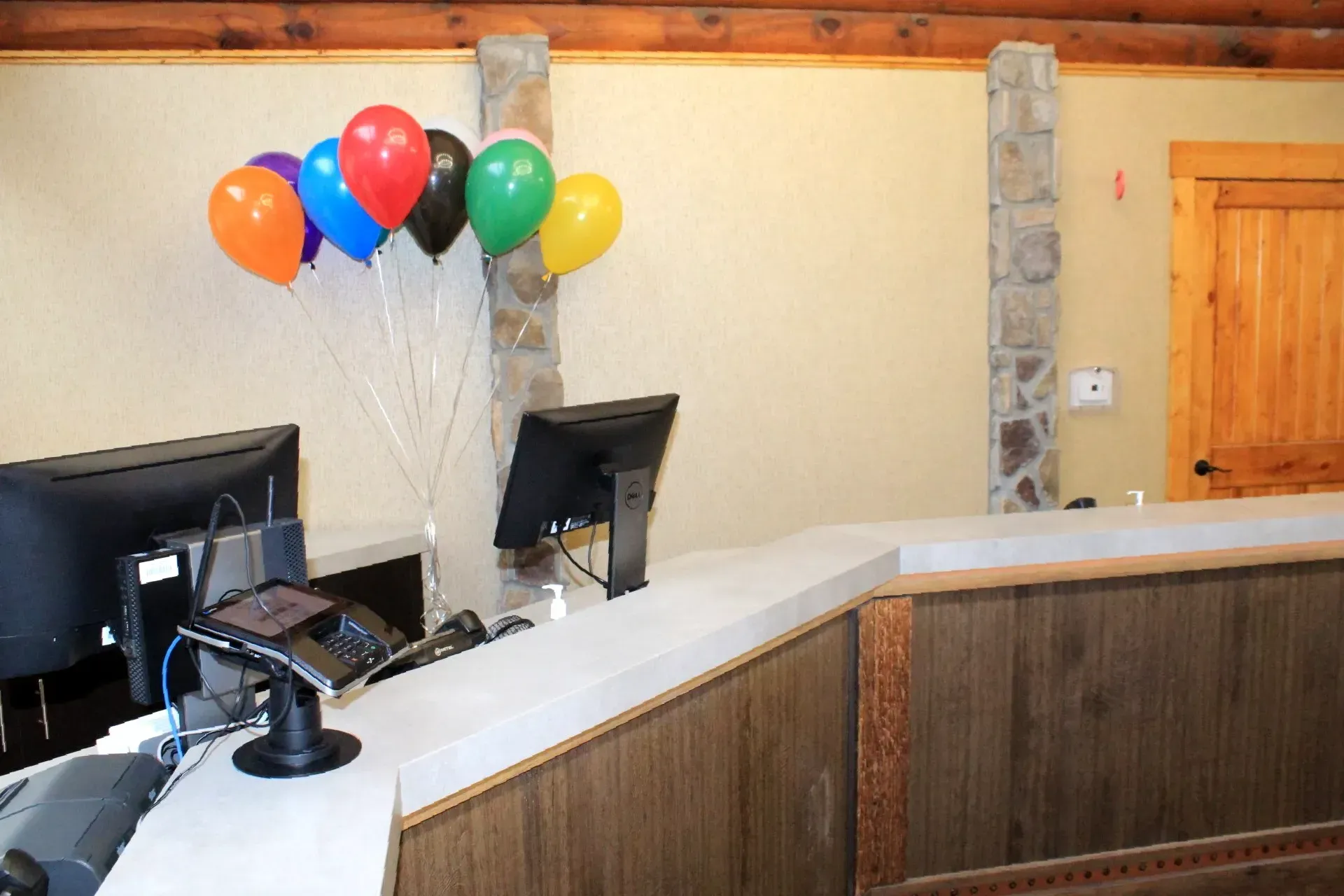 Reception desk with balloons; monitors and phone on countertop. Wooden accents and beige walls.