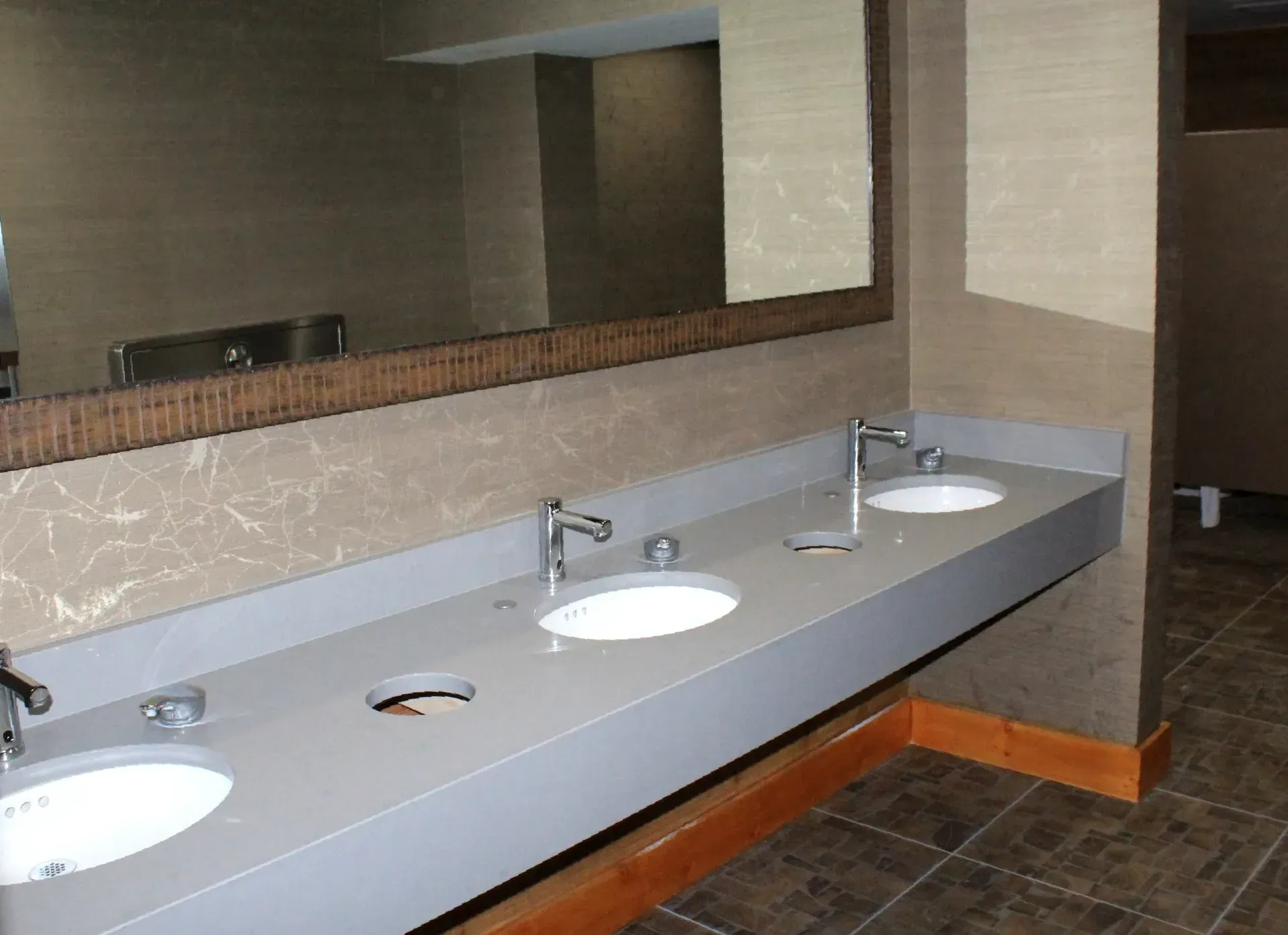 Public restroom with three sinks and a large mirror. Gray countertops, brown accents, and beige patterned walls.