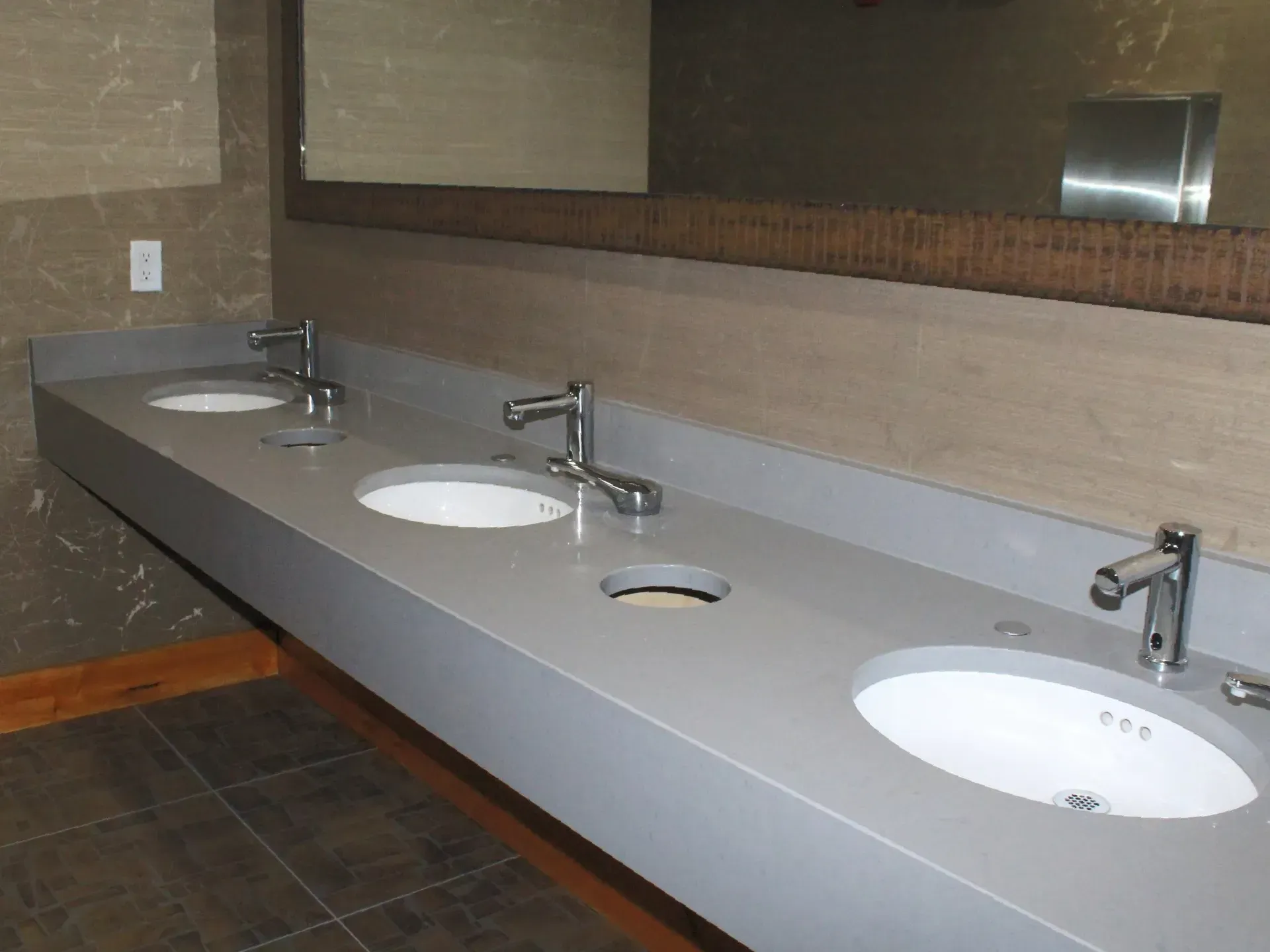 Public restroom with three sinks and chrome faucets against a gray countertop and a large mirror.