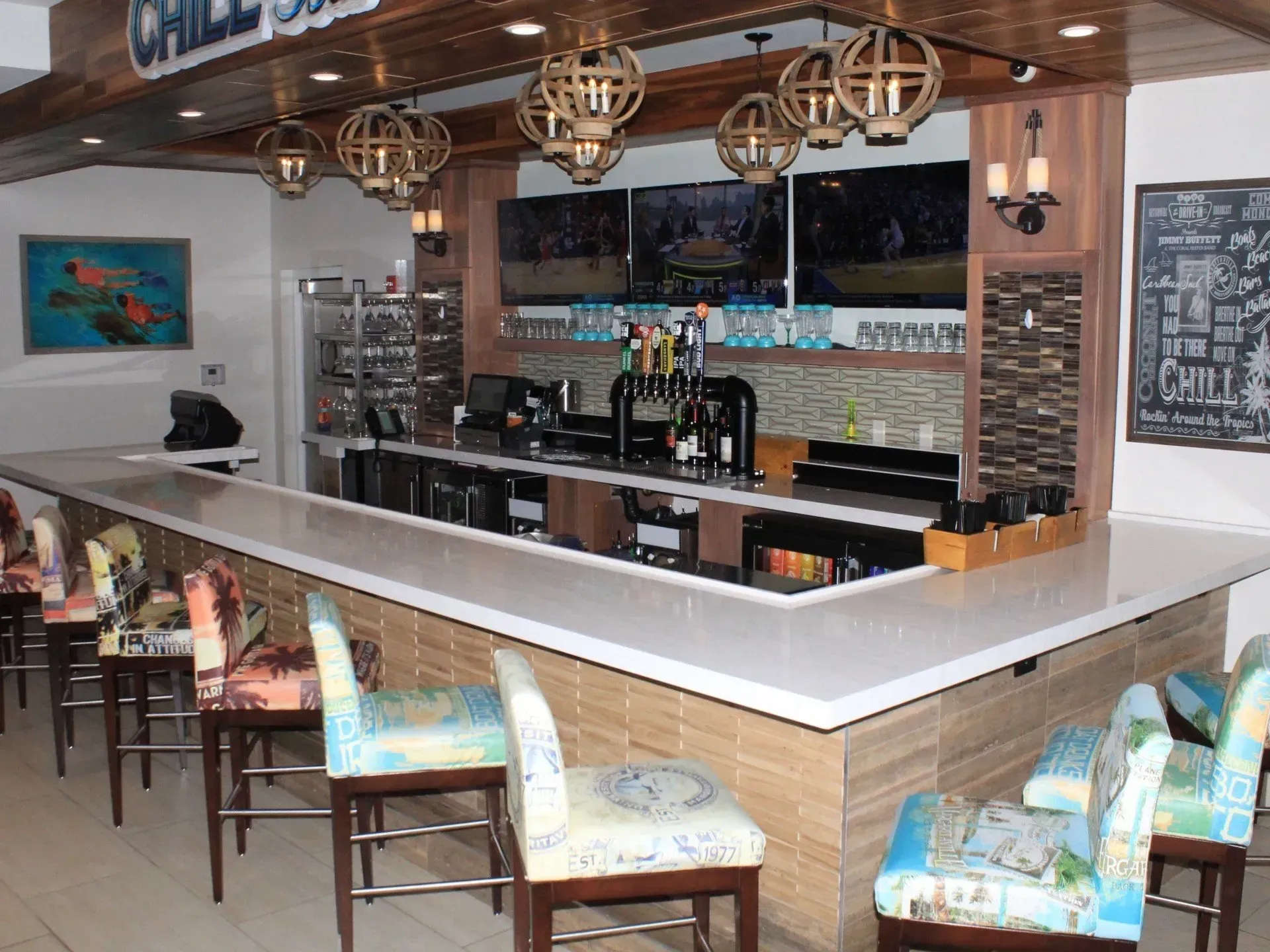 Bar with white countertop, stools with colorful fabric, televisions above bar, and hanging globe lights.
