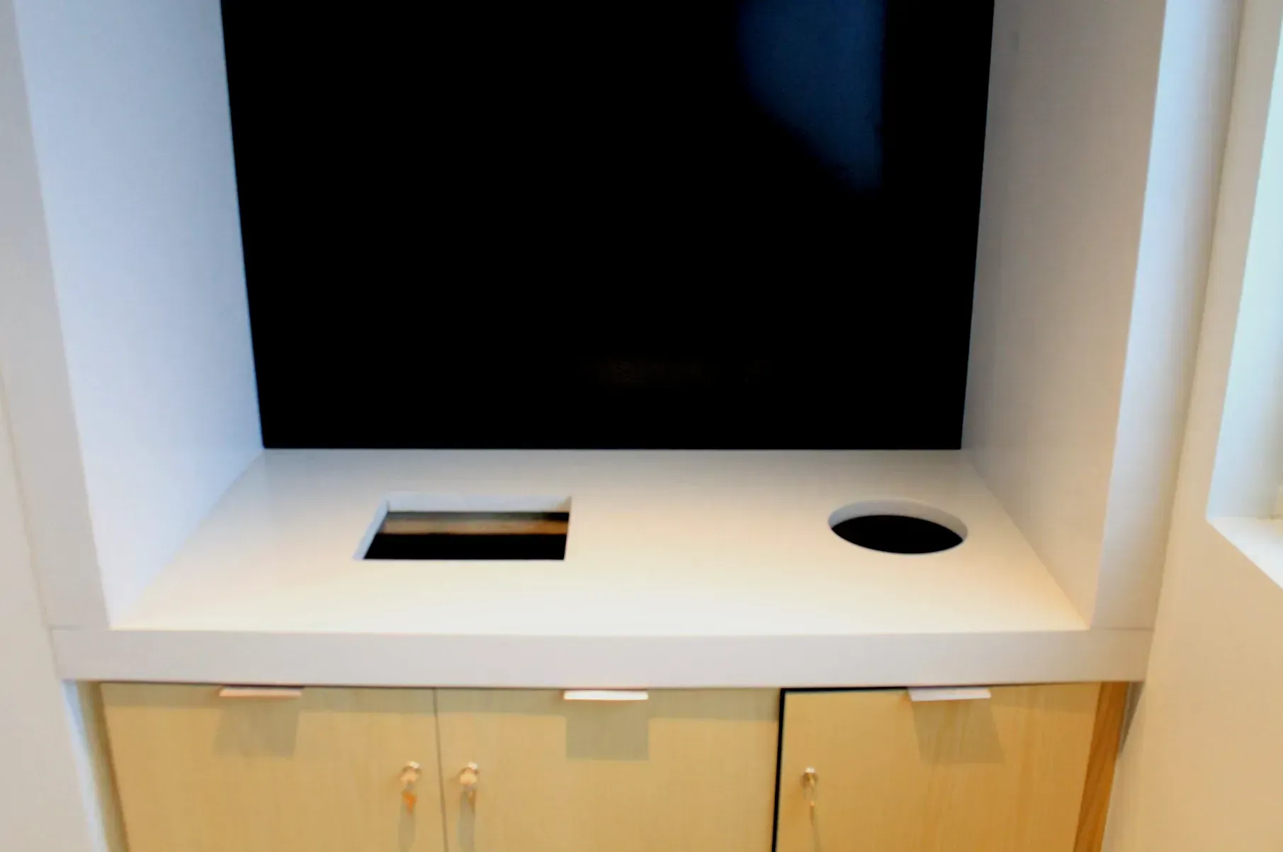 White-walled built-in entertainment center with a black TV, light wood cabinets, and two holes in the countertop.
