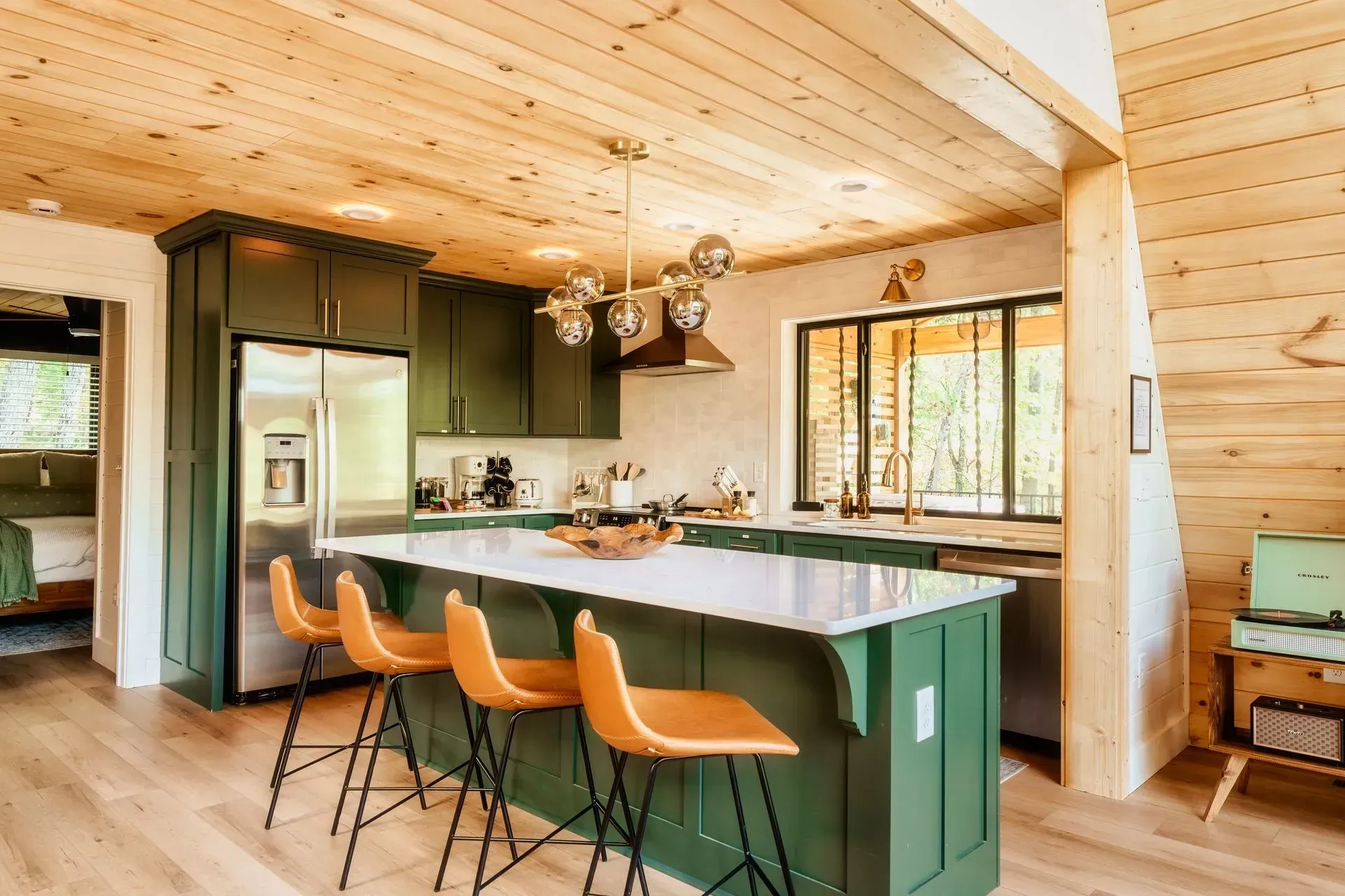 Modern kitchen with green island, orange bar stools, wood-paneled walls, and large windows