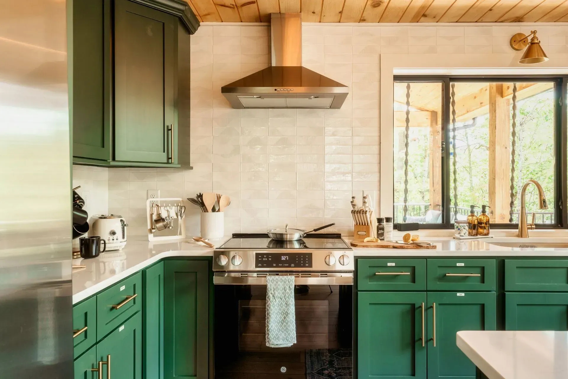 Green kitchen with white tile backsplash, stainless range hood, and sunlight through a window over the sink