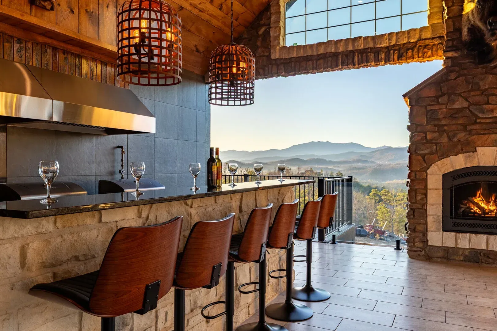 Outdoor bar with mountain view, stone facade, stainless steel hood, seating, fireplace, pendant lights.