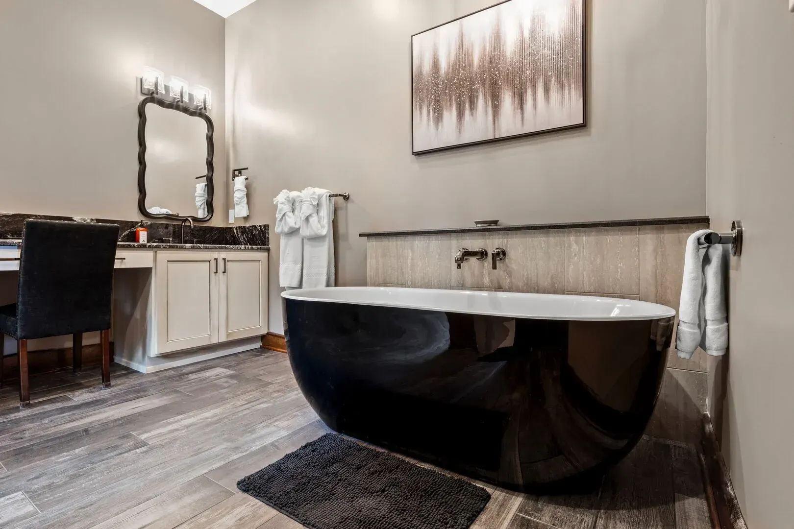 Bathroom with a black bathtub, vanity, mirror, and artwork. Wooden floor, neutral walls, and a towel rack.