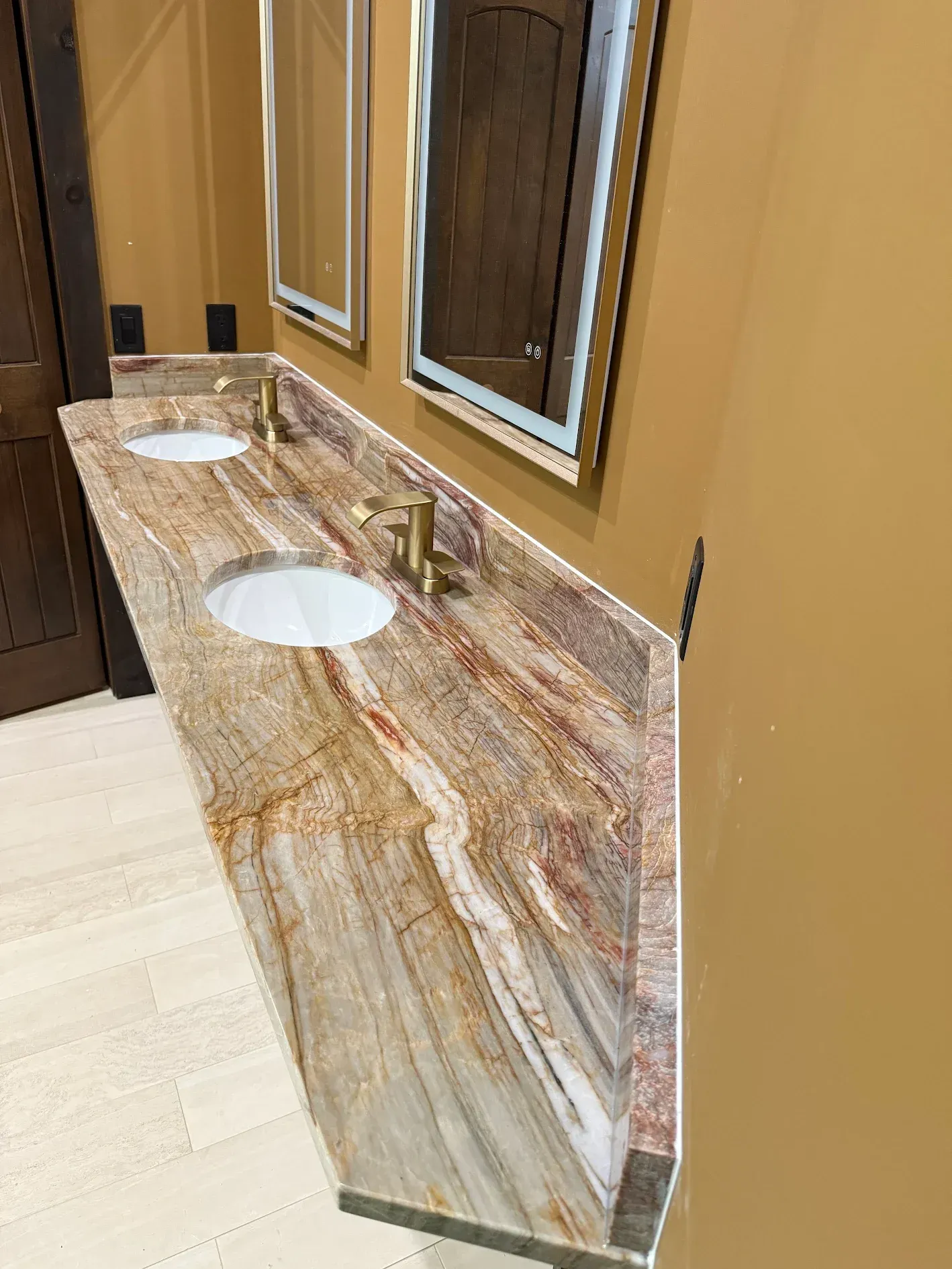 Granite countertop with two sinks and gold faucets in a bathroom, with mirrors and a wooden door.