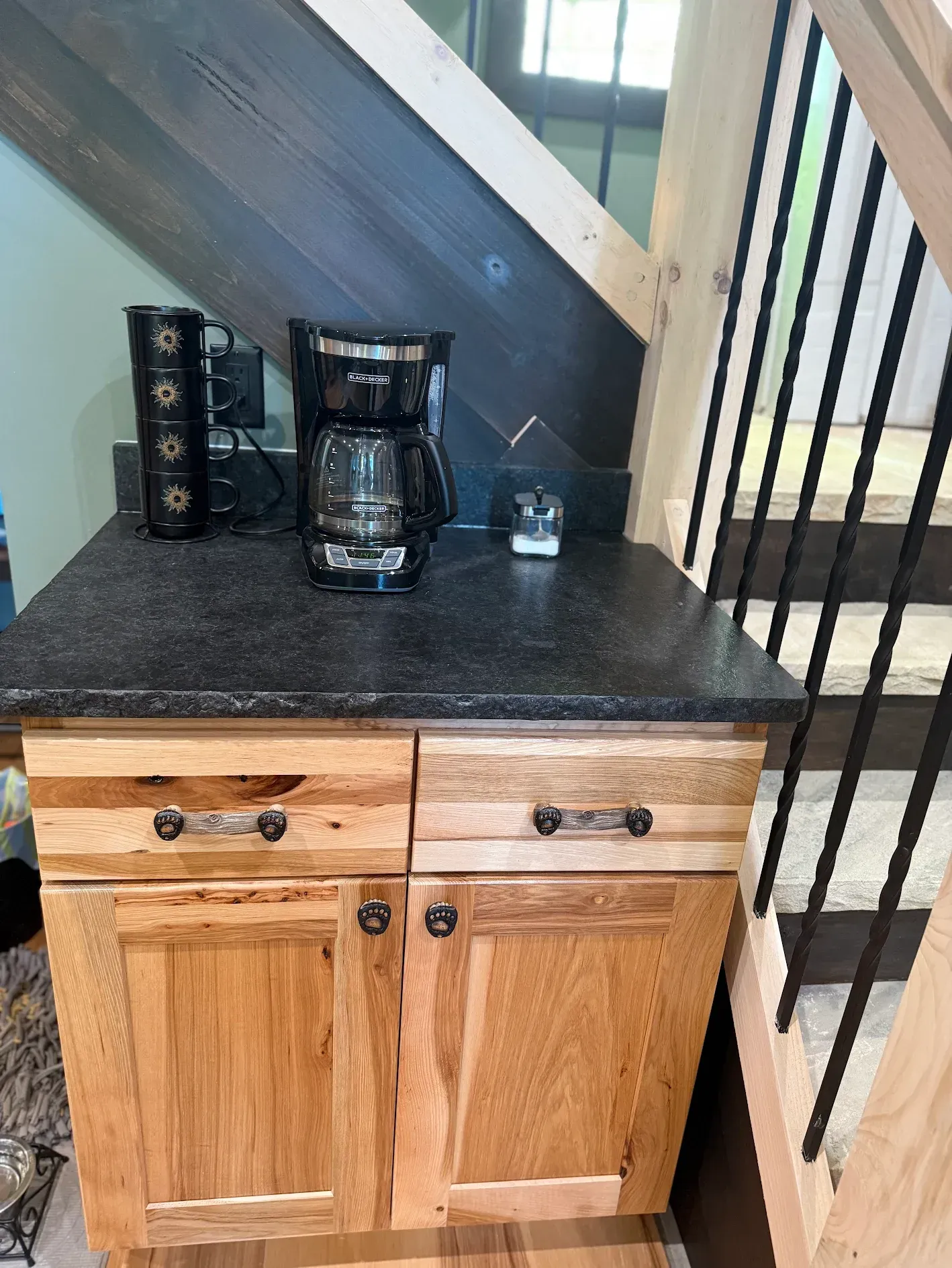 Coffee station with coffeemaker, under wooden stairs. Cabinet is natural wood with dark countertop.