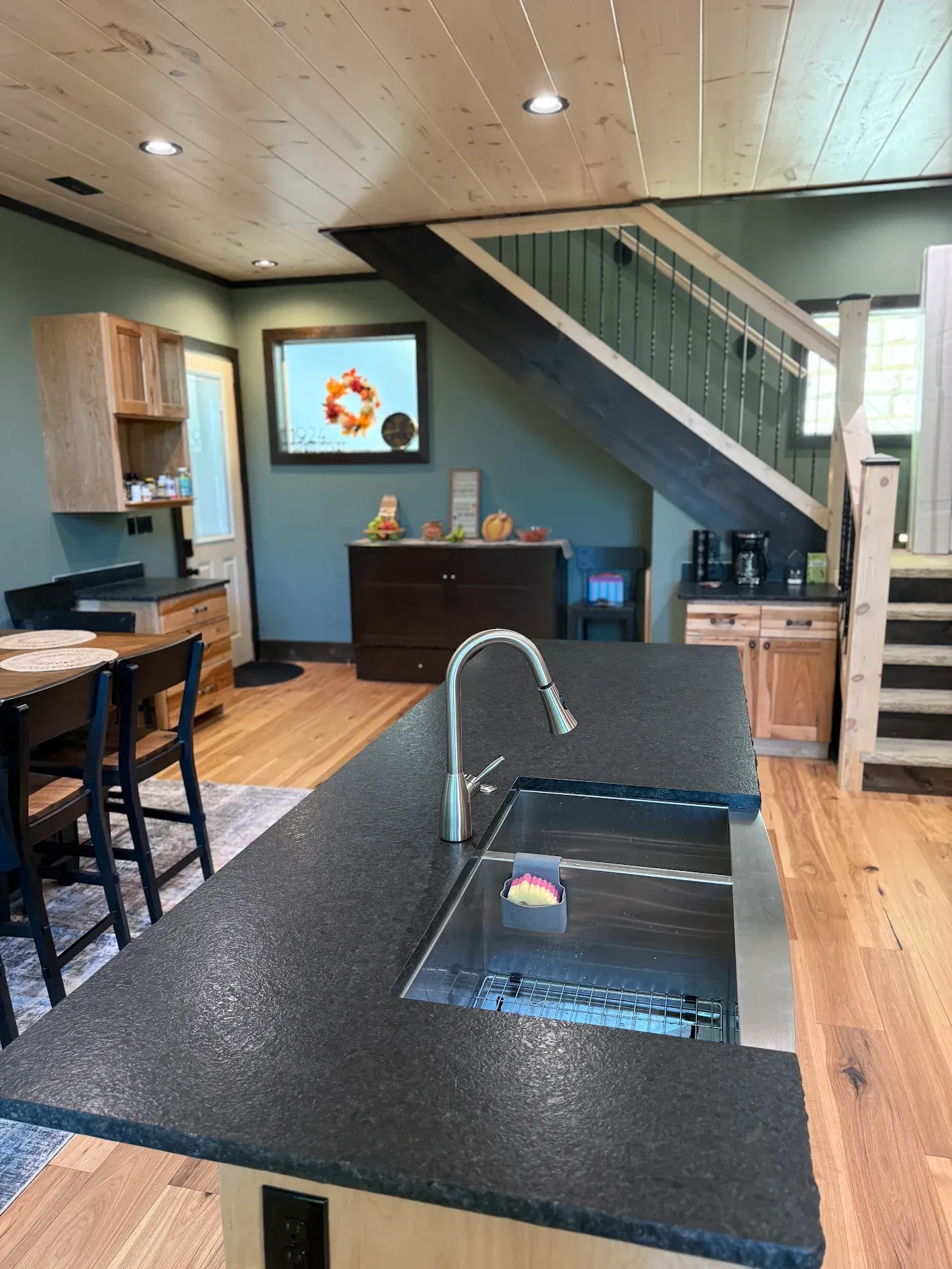 Kitchen with island sink, stairs, and wooden cabinets. Dark countertop, sage green walls, light wood floor.