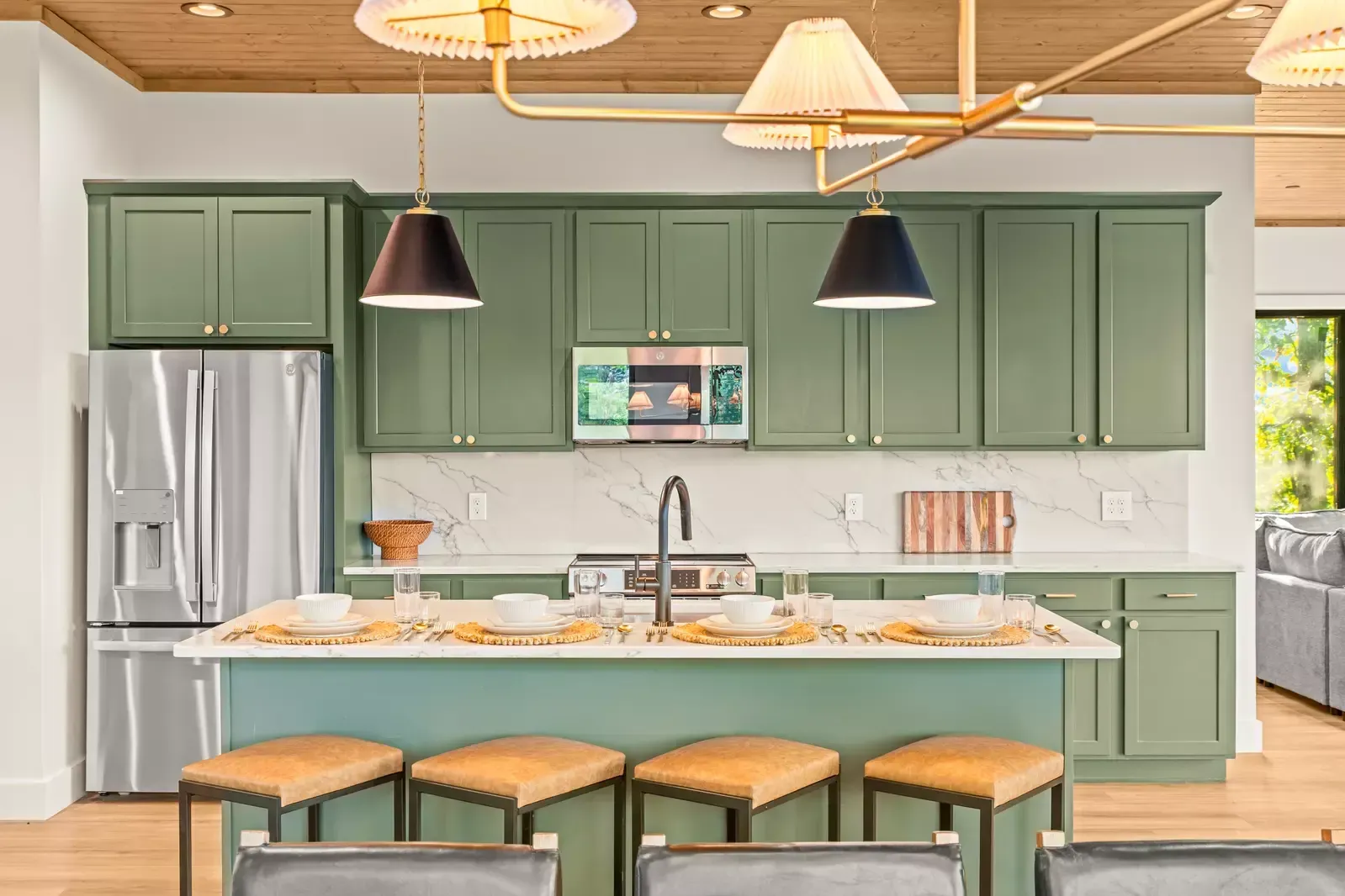 Green kitchen with island, pendant lights, stainless fridge, and four woven stools