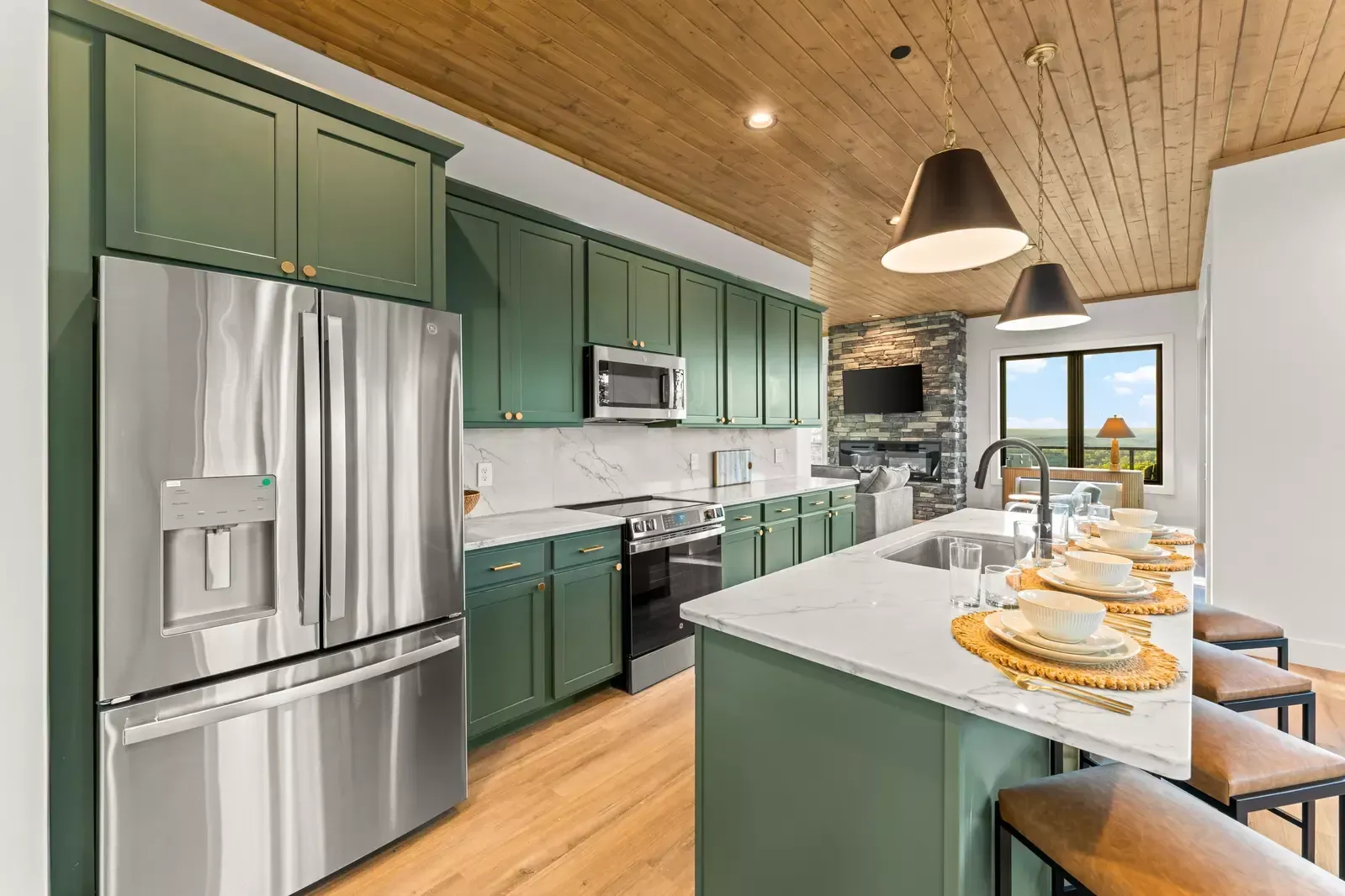 Modern green kitchen with stainless steel appliances, island seating, and a dining table set near a window.