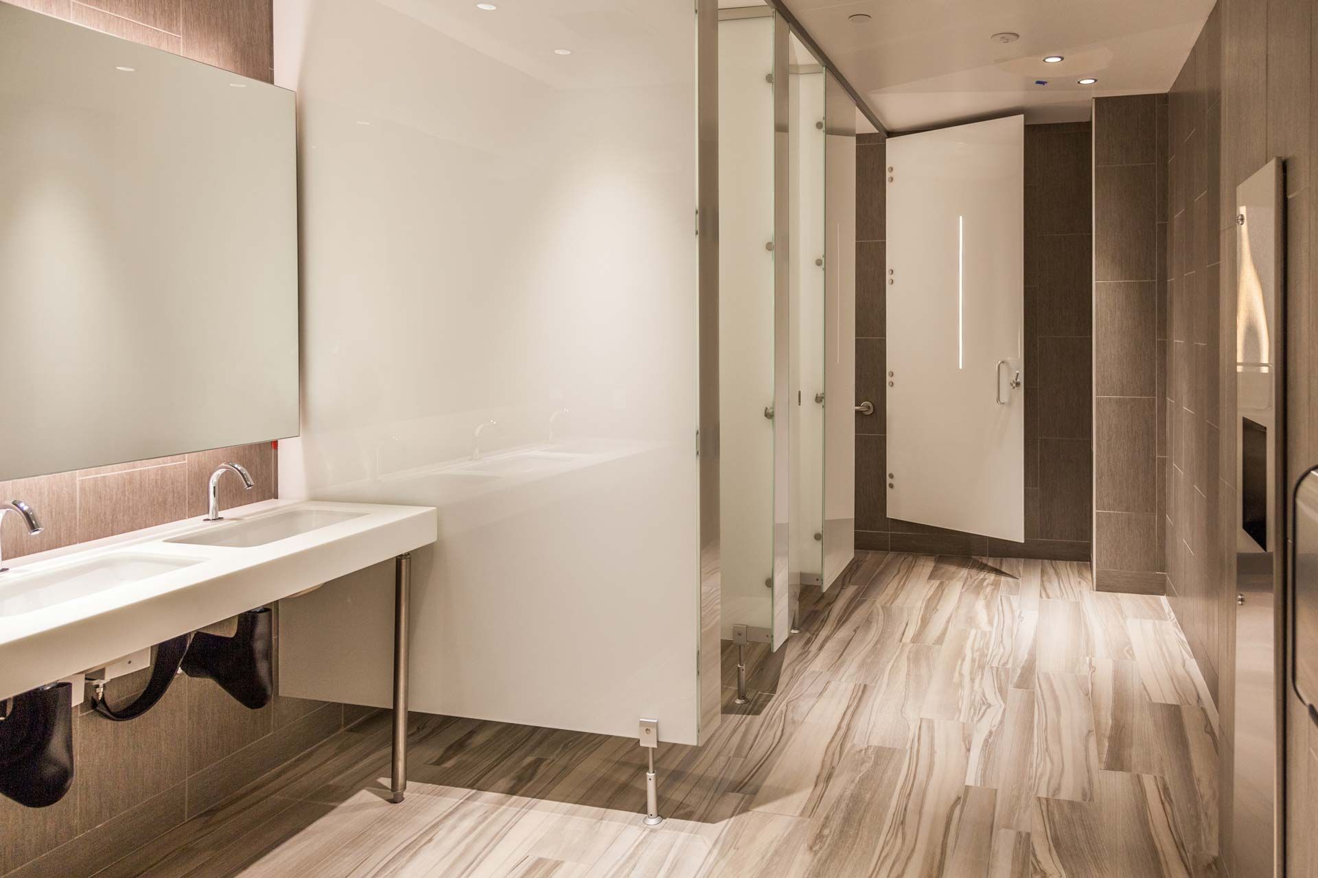 Modern restroom with long sink, stalls, mirrors, and wood-patterned floor under warm lighting