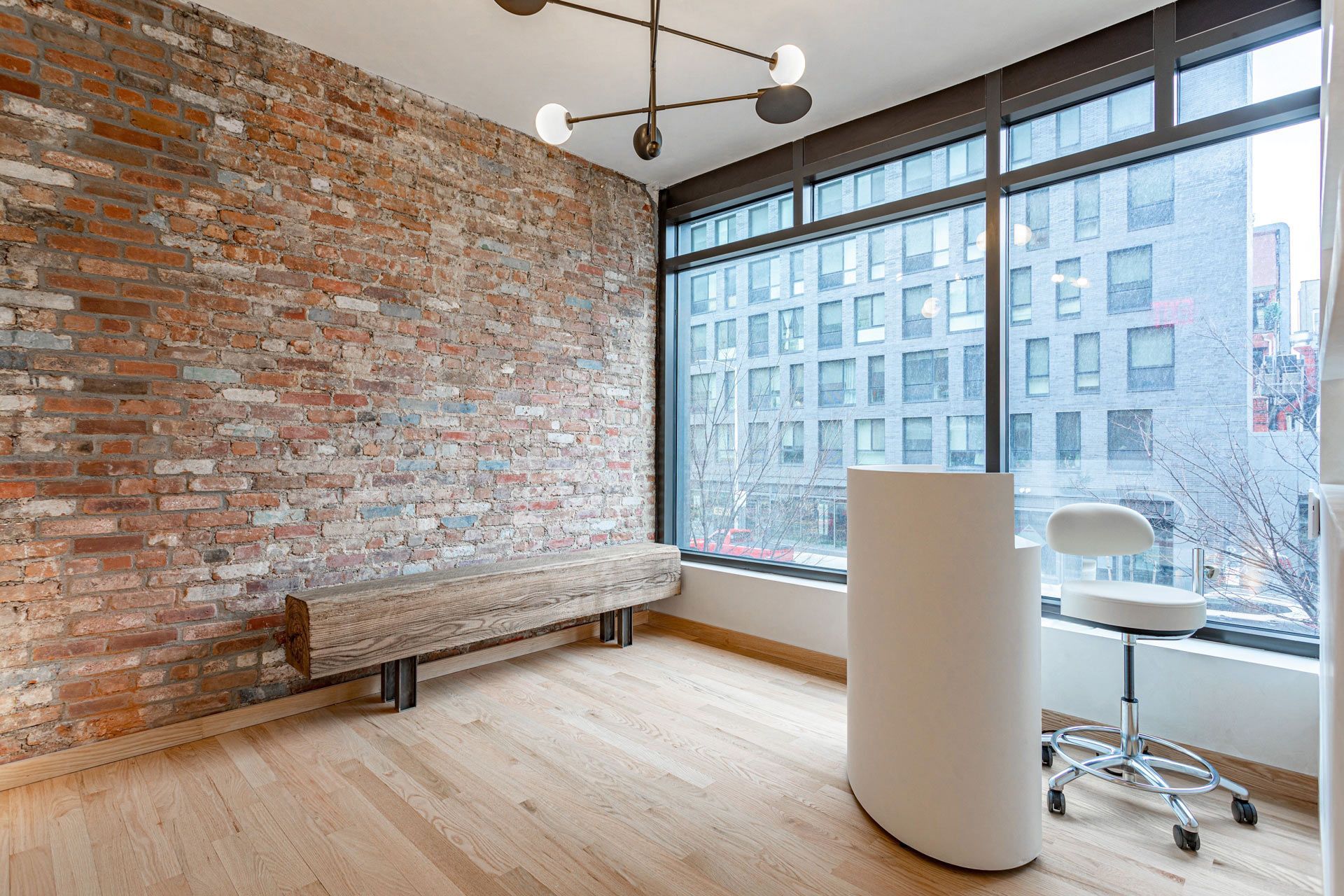 Modern office room with exposed brick wall, large windows, wood floor, and minimalist table and chairs