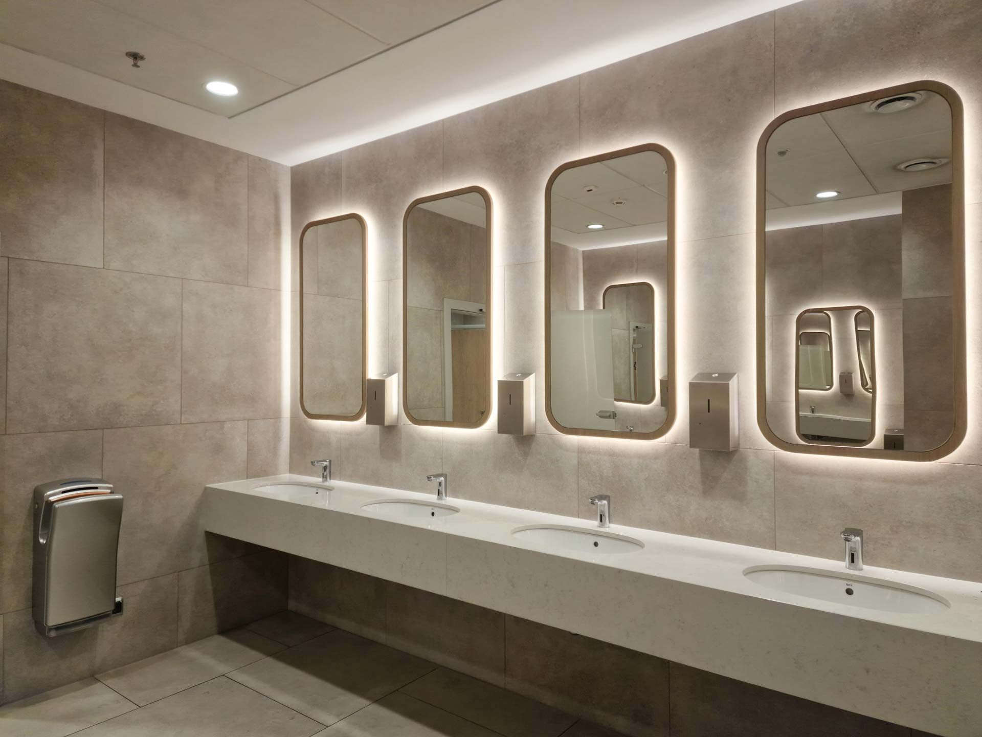 Modern public restroom with long white sink counter and illuminated mirrors on beige tiled walls.