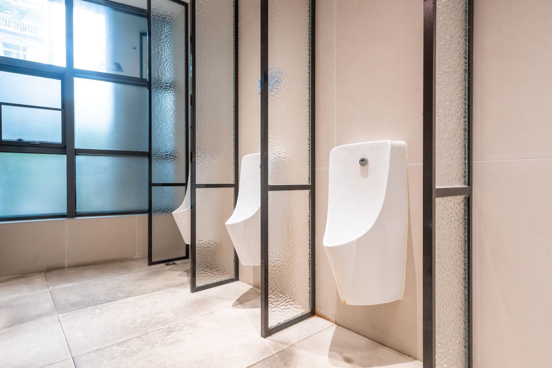 Modern restroom with white urinals, black dividers, and a large frosted window in soft daylight