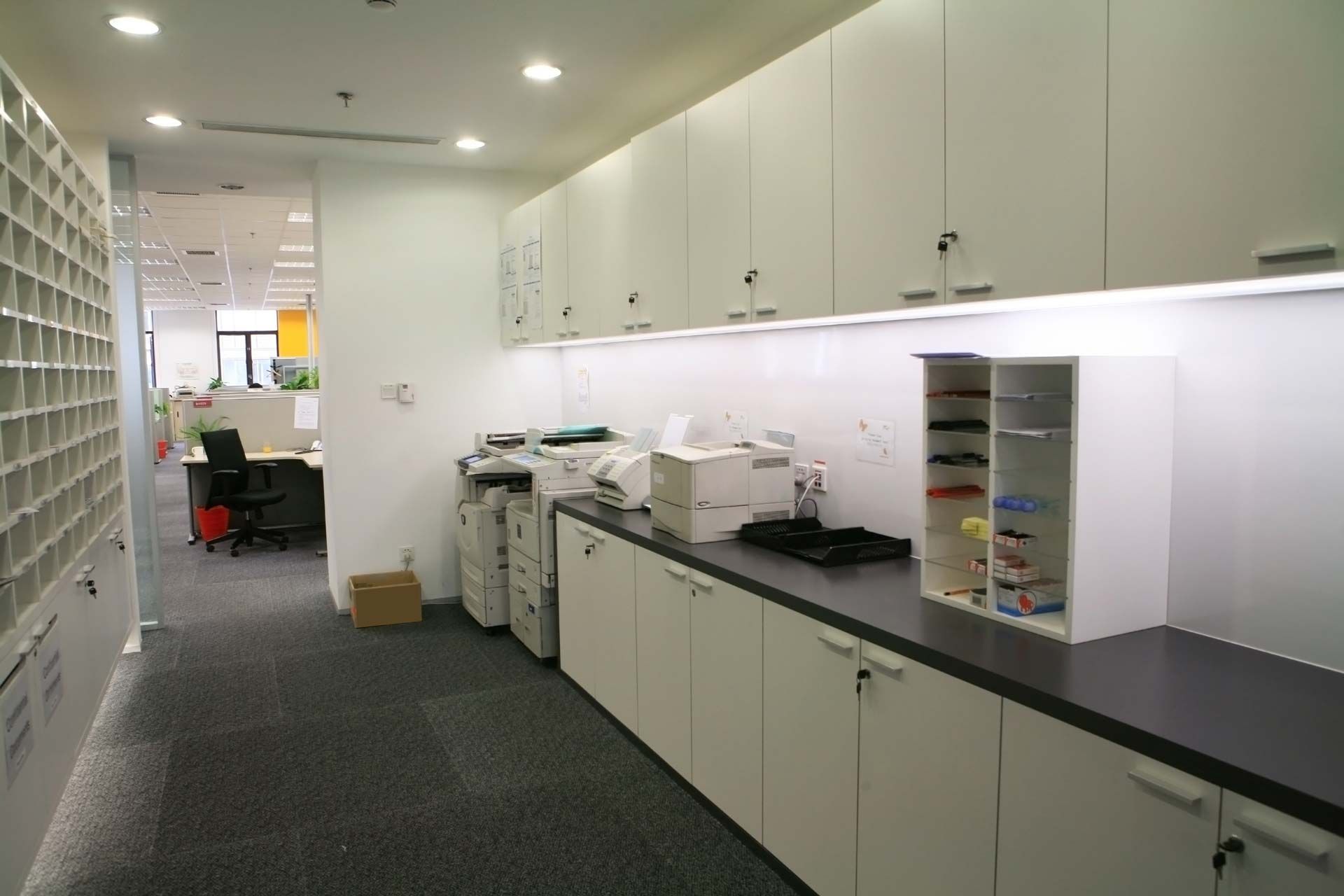Office supply room with white cabinets, black countertop, copy machines, and shelves along a narrow hallway