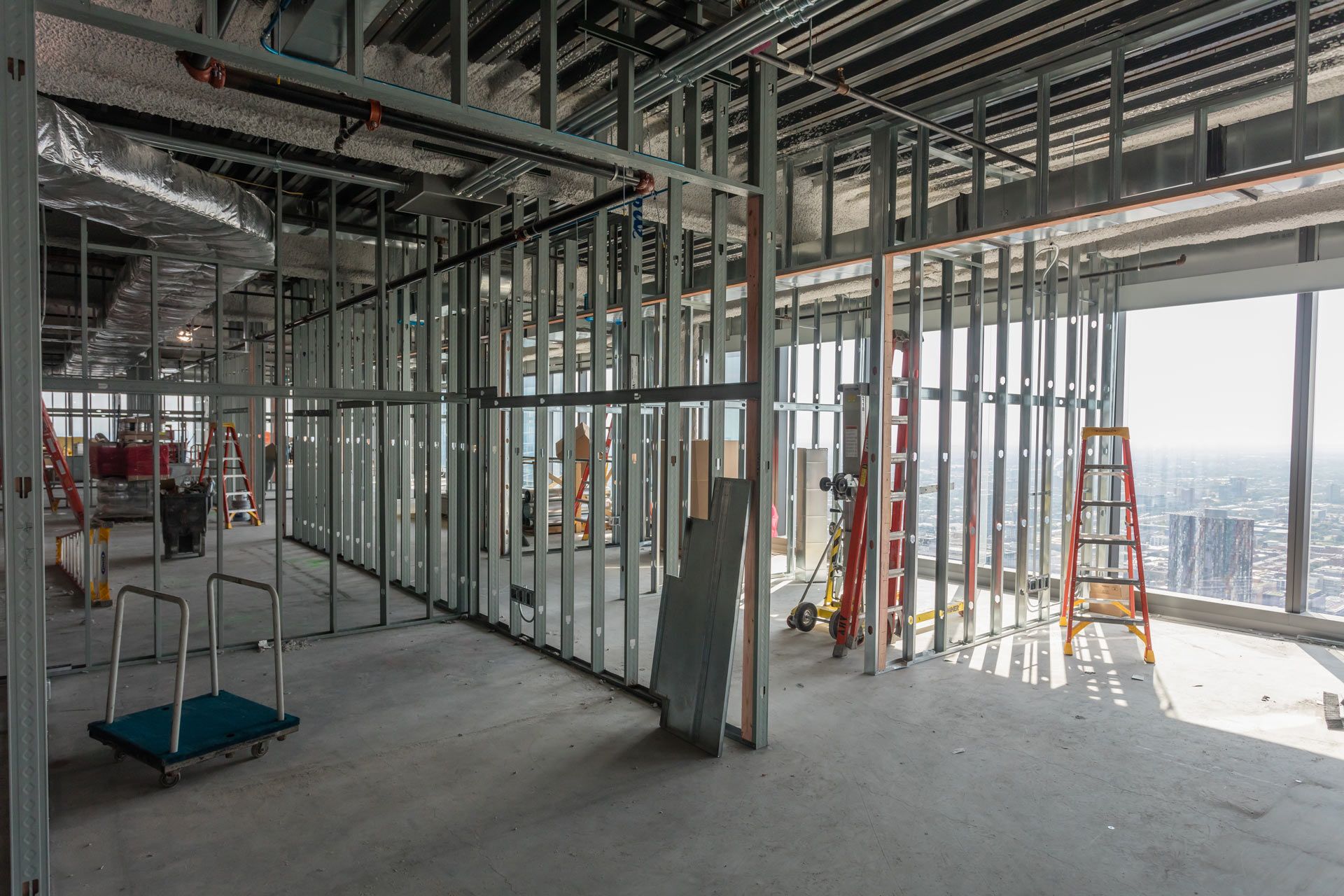 Interior of a building under construction with exposed framing, ladders, and sunlight through large windows.