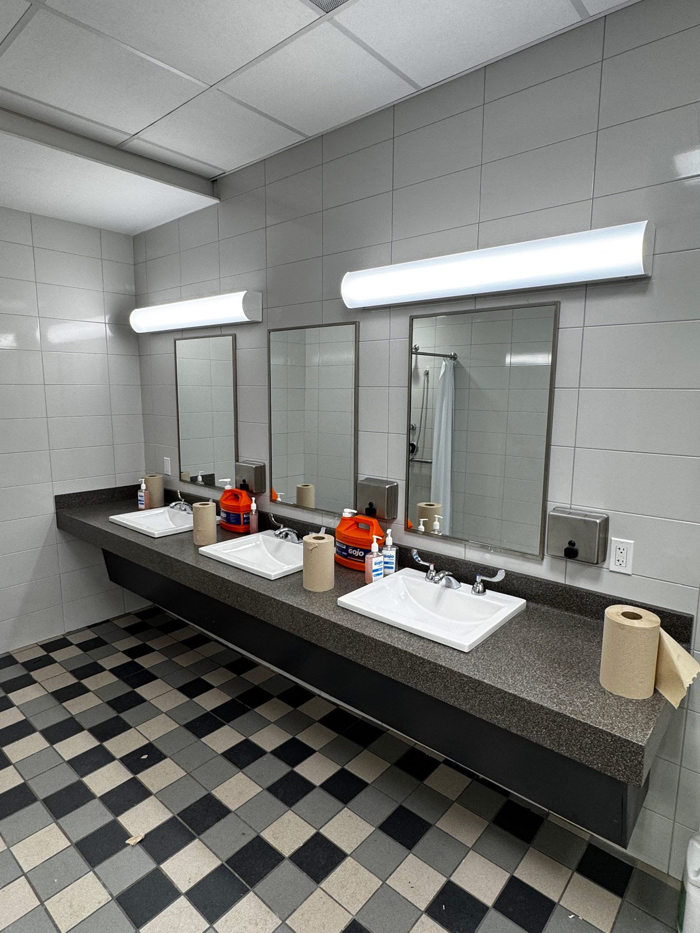 Public restroom sink area with three mirrors, bright lights, checkered floor, and paper towels on the counter