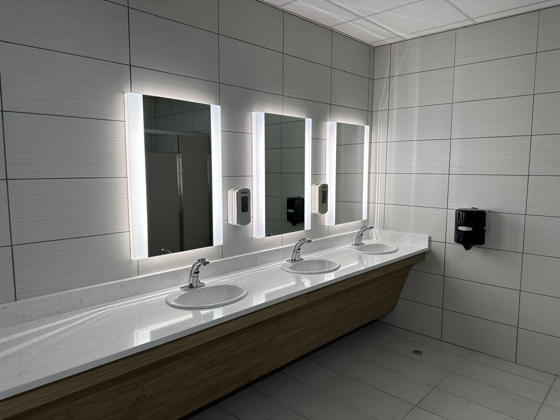 Modern restroom with three lit mirrors, white sinks, and a long wooden vanity against tiled walls.