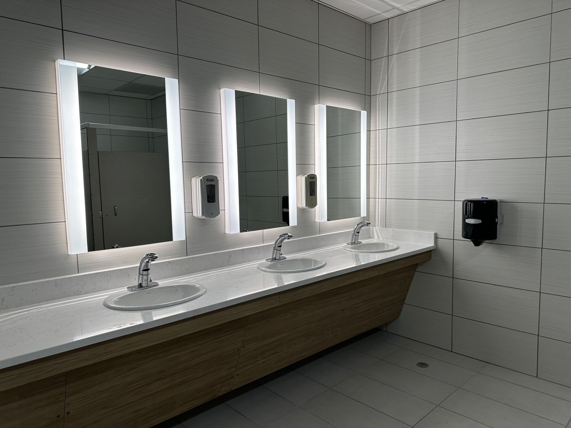 Accessible restroom with three lit mirrors, long sink counter, and soap dispensers on tiled walls.