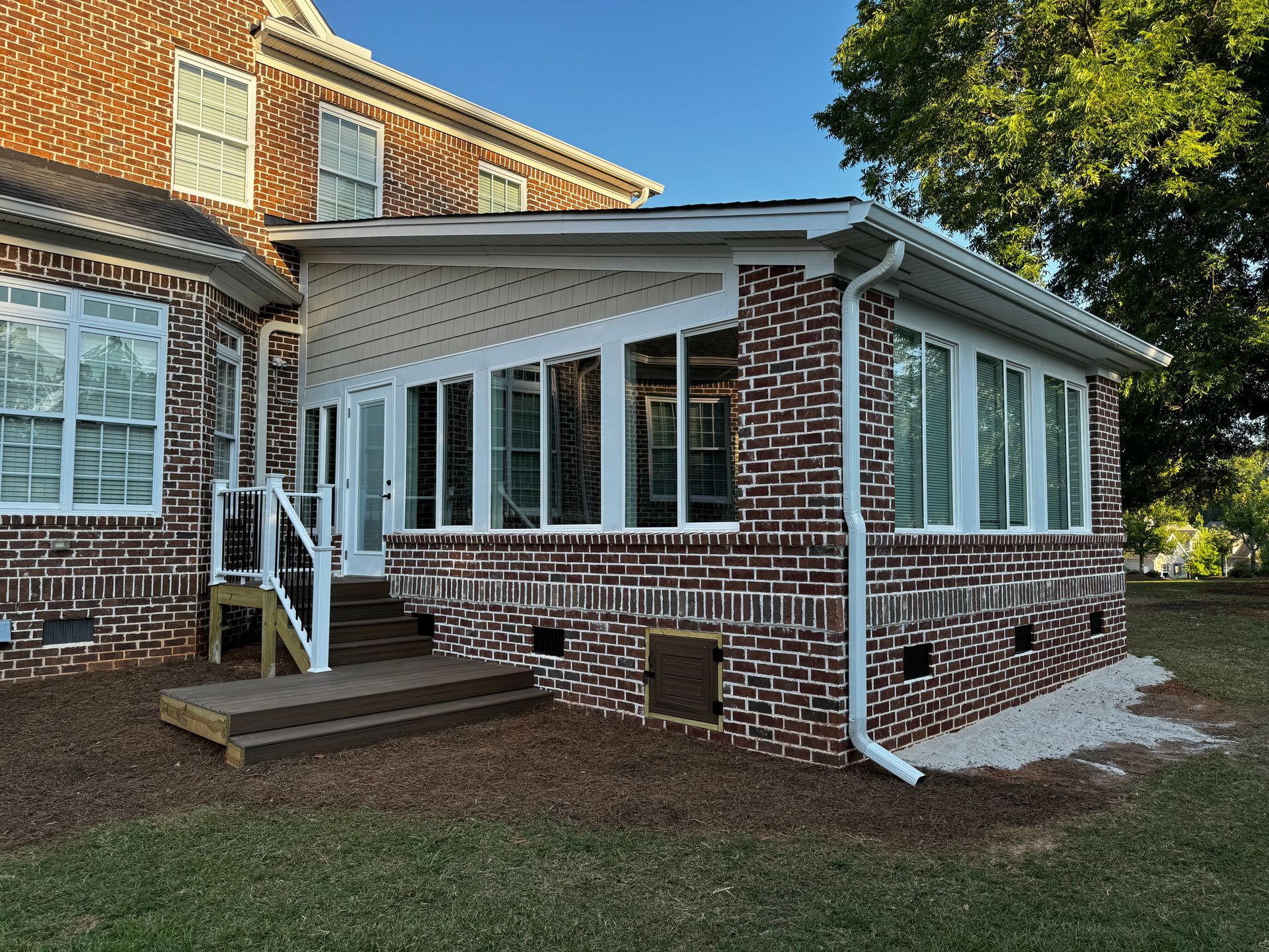 Brick house sunroom with white-framed windows, porch steps, and a downspout in the yard.