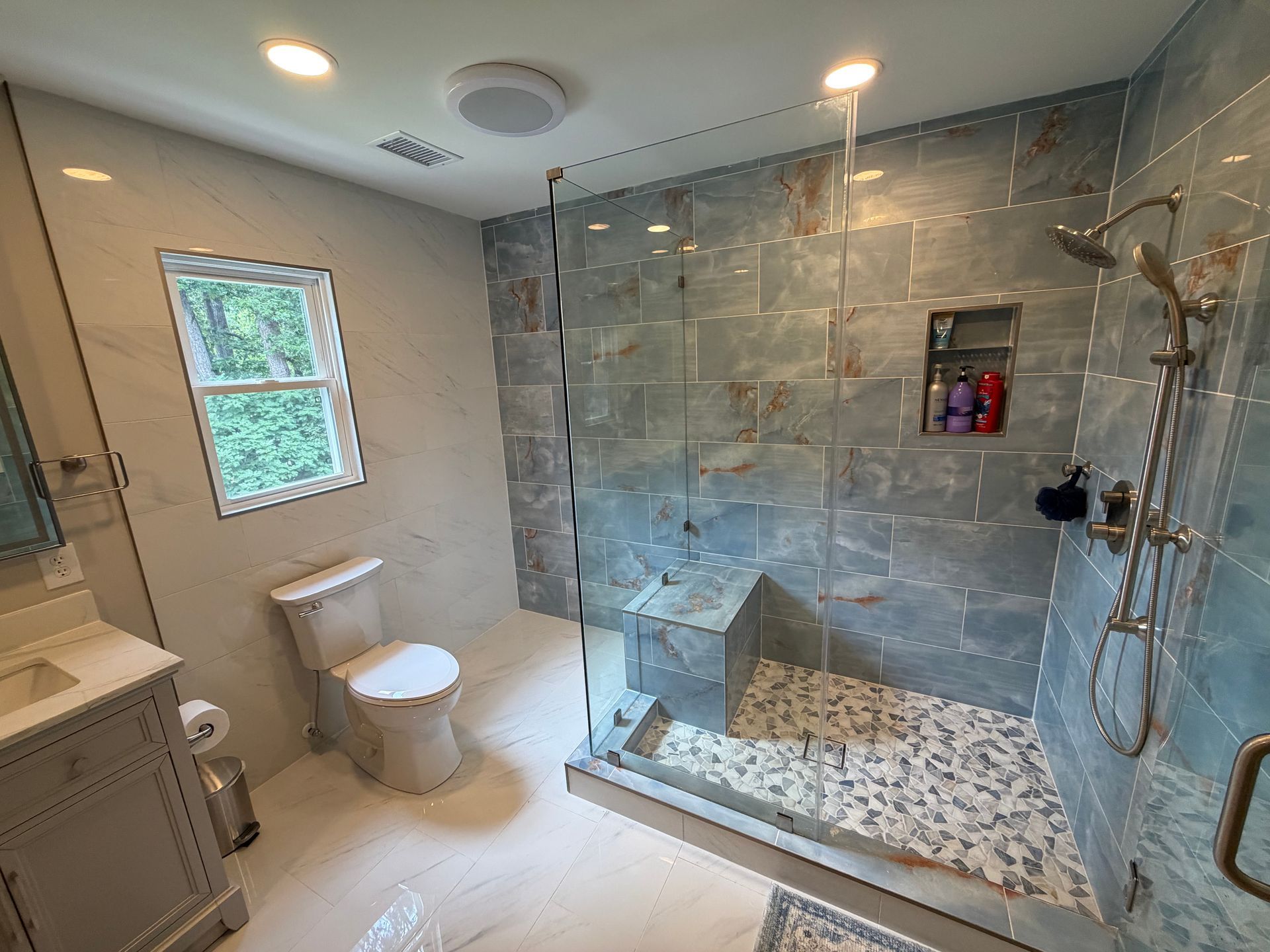 Modern bathroom with tiled walk-in shower, toilet, vanity, and window in soft gray tones