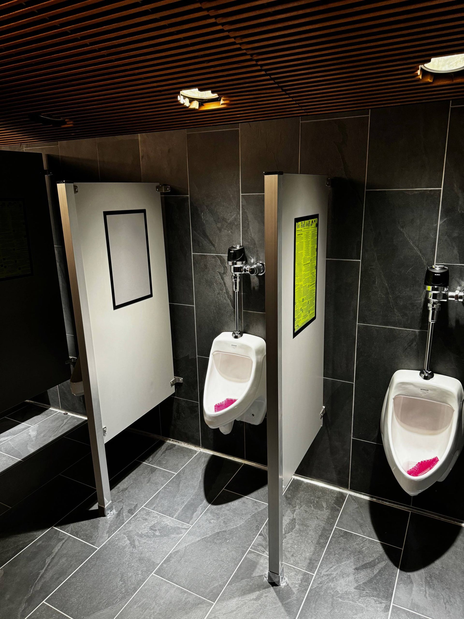 Public restroom with black tile, two urinals, and white stall doors with yellow signs.