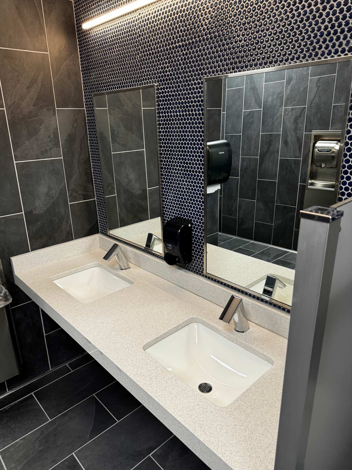 Modern restroom with double sinks, large mirrors, black tile, and speckled white countertop