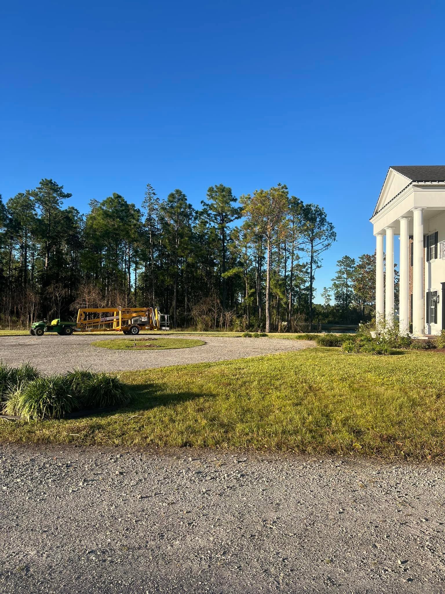 Gravel courtyard with a white-columned building, pine trees, and a yellow construction vehicle under a clear blue sky