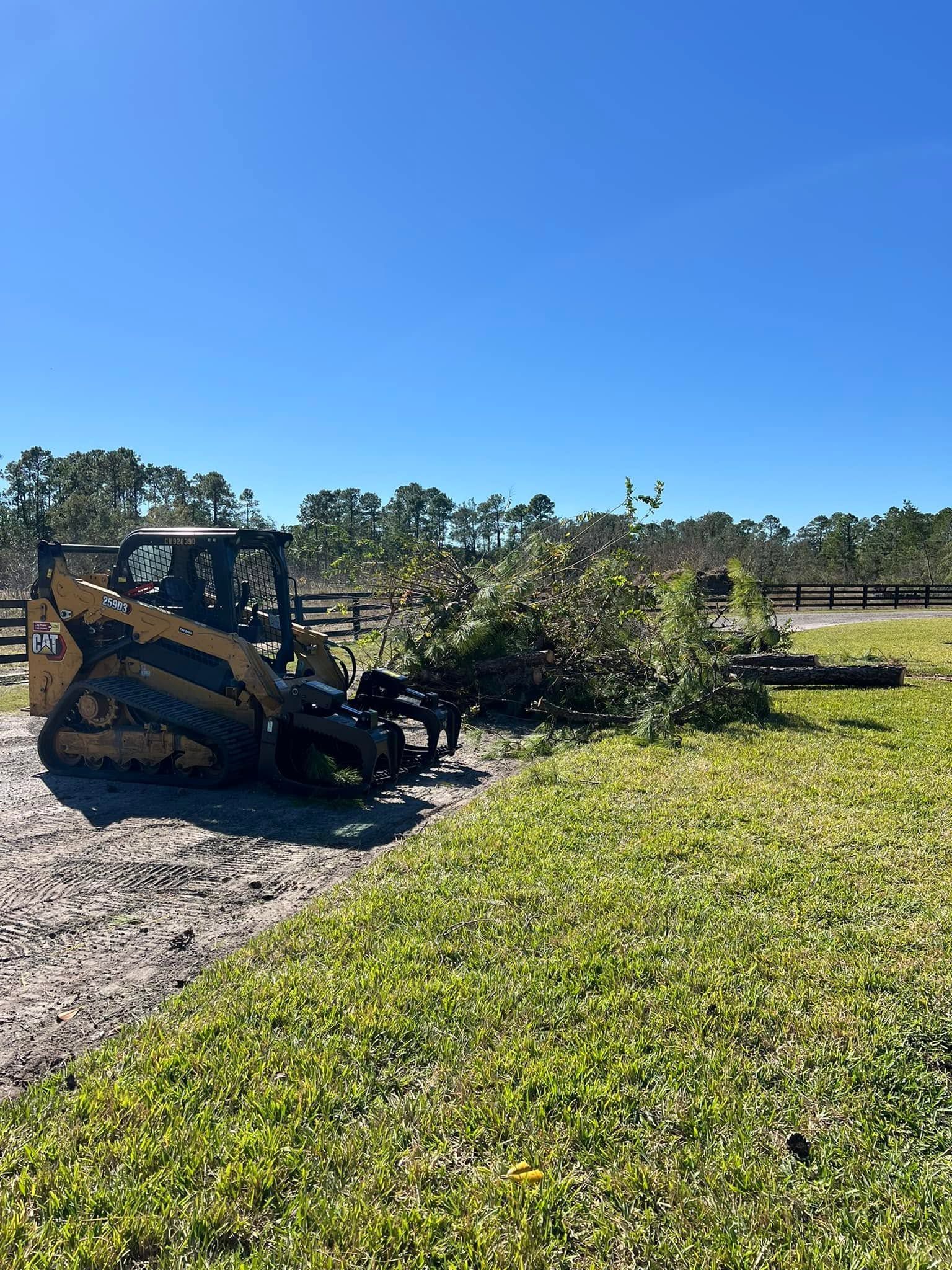 A yellow skid steer works beside a gravel path and green field under a clear blue sky.