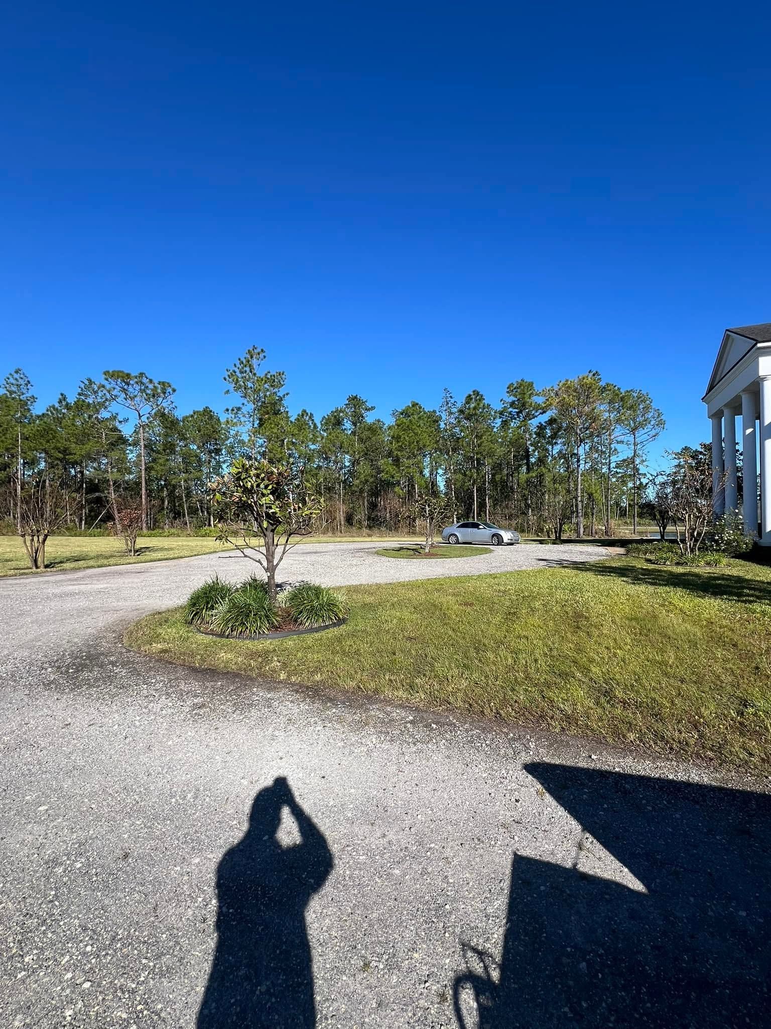 Gravel driveway beside a white house under a bright blue sky, with trees and a small car in the distance.