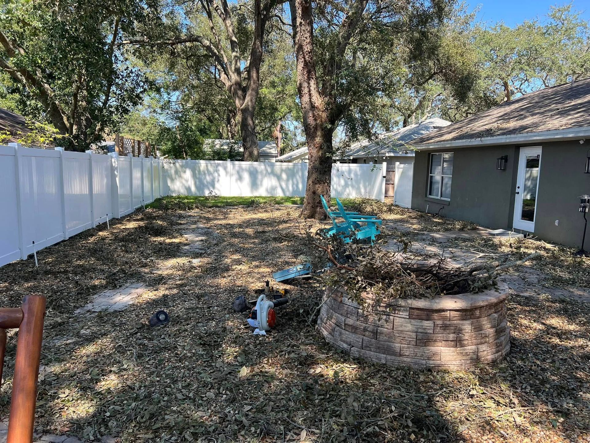 Backyard with large trees, white fence, gray house, and a brick fire pit amid leaf-covered ground
