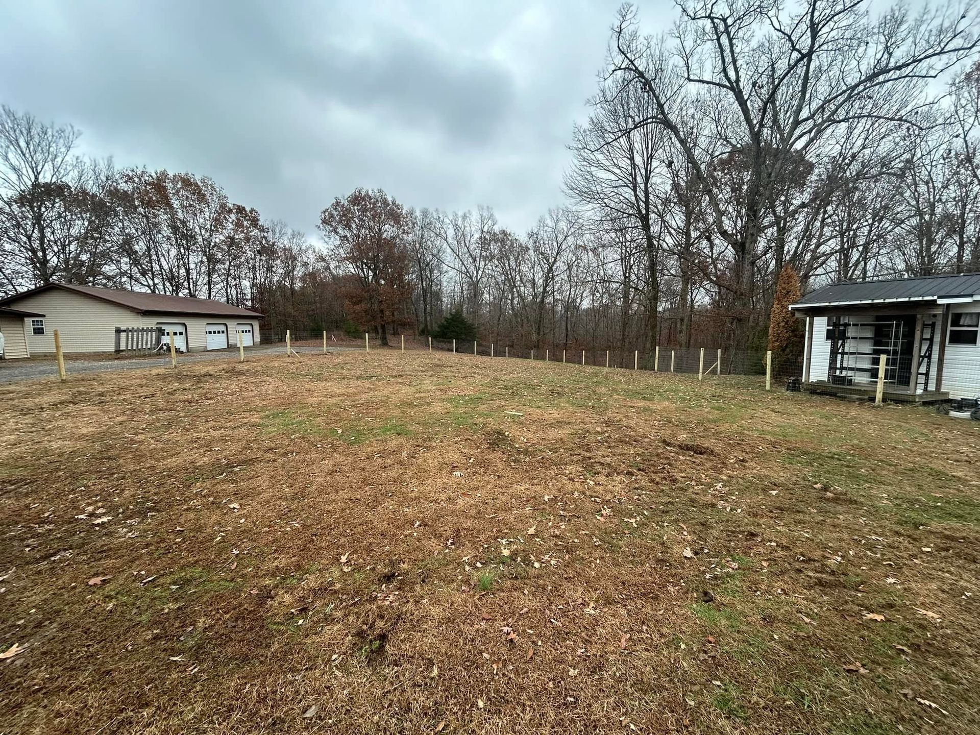 Empty grassy lot with bare trees, a house on the left, and a porch on the right under a cloudy sky