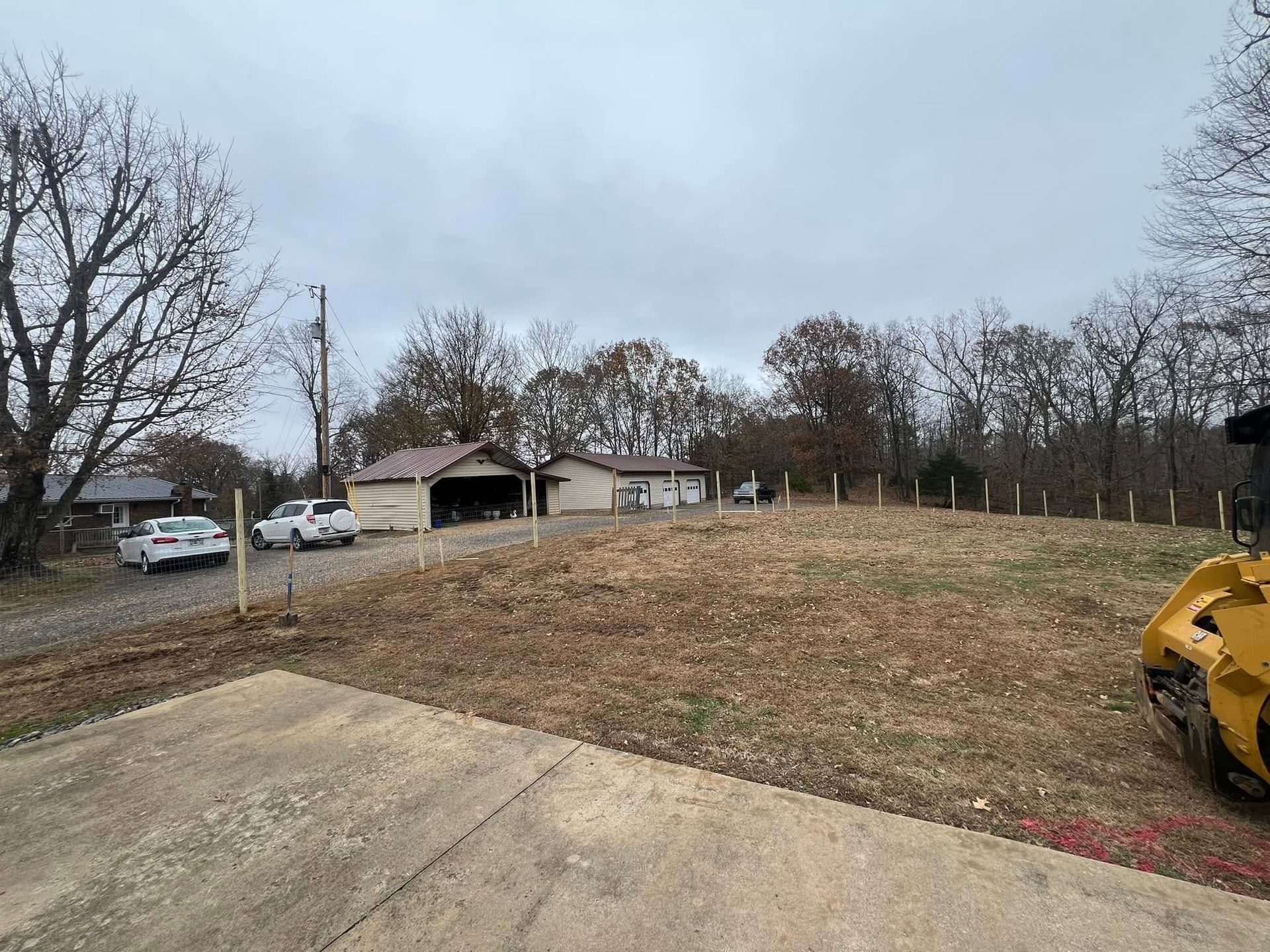 Gravel lot beside a grassy field with parked cars, a barn, leafless trees, and a yellow bulldozer edge