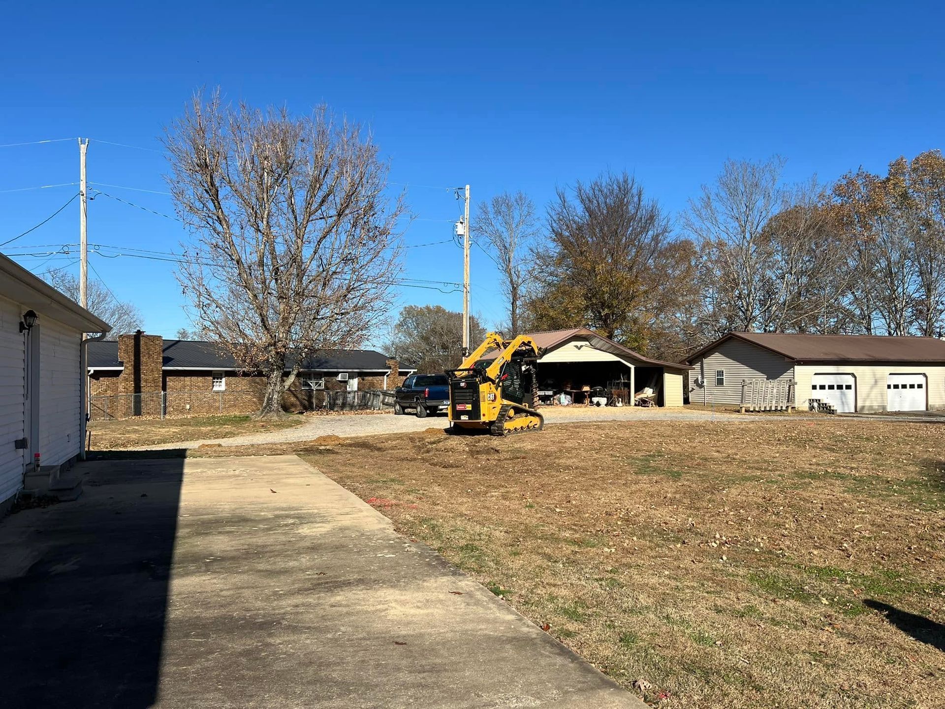 Backyard with a yellow excavator near a house, beside a driveway and grassy yard under a clear blue sky
