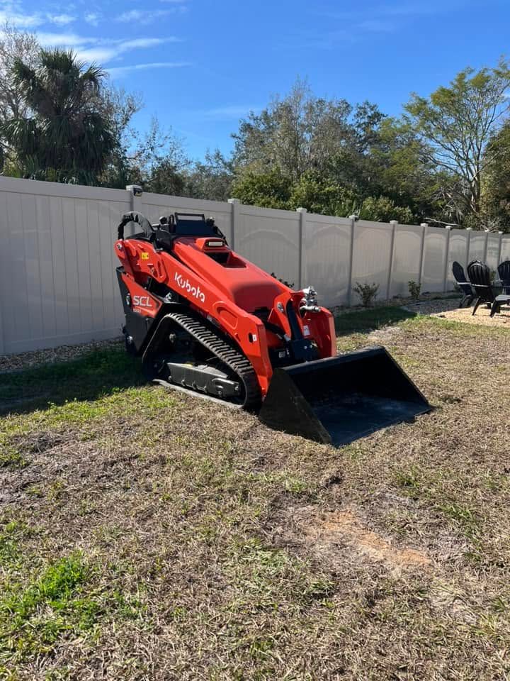 Red compact loader with black bucket on grass beside a white fence under a blue sky