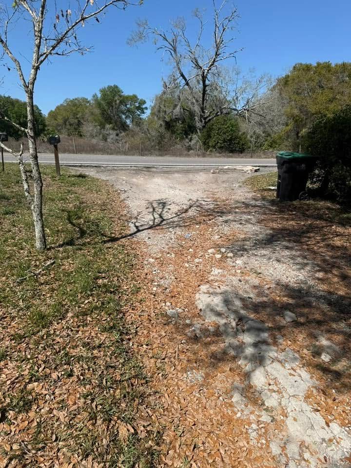 Gravel driveway leading to a road, with trees, dry leaves, and a green trash bin on the right.