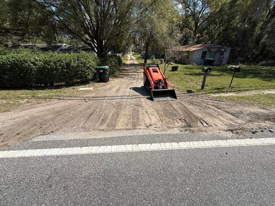 Orange skid steer parked on a dirt driveway beside a road, with trees and a house in the background