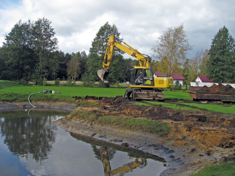 Yellow excavator digging by a pond on a muddy construction site with trees and houses in the background
