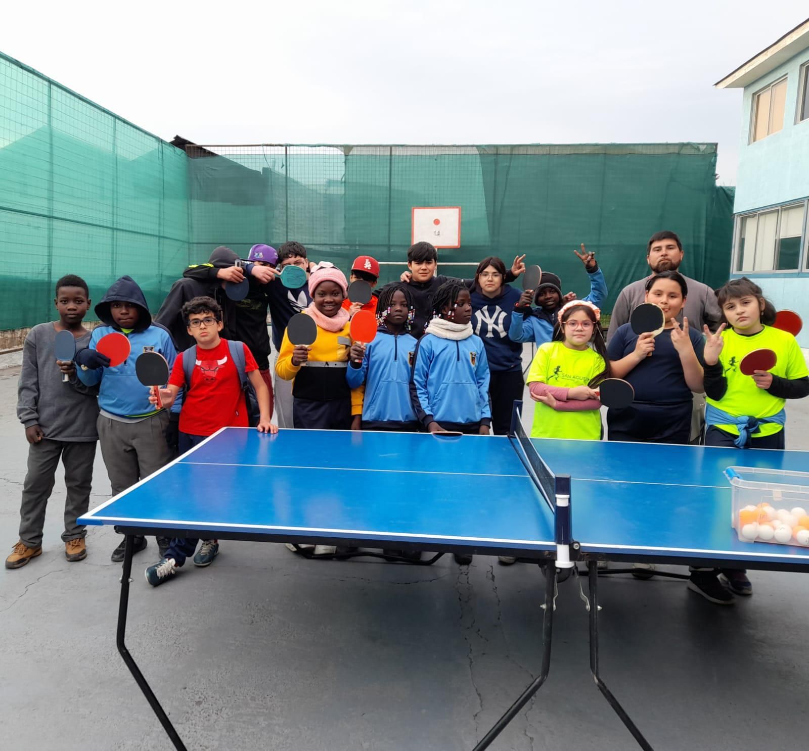 Grupo de niños con raquetas de ping pong posando junto a una mesa azul al aire libre. Algunos las sostienen.